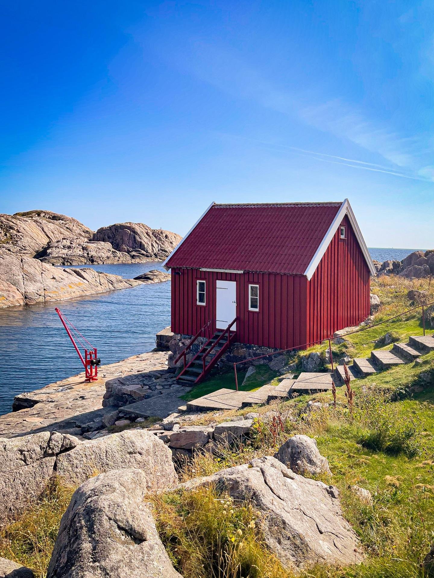 Red wooden house with a white door and trim located on a rocky coastal landscape with sparse greenery. Steps lead up to the house and a red crane stands on a small pier beside it. Clear blue sky overhead and rocky hills in the background.