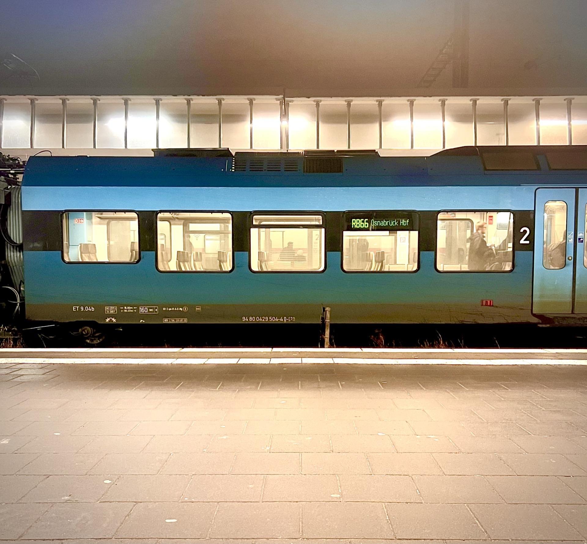 Blue commuter train stopped at a platform at night, labeled as "R865 Innsbruck Hbf." The illuminated interior reveals empty seats and a few silhouetted passengers. A platform edge and a lit station roof overhang are visible above the train.