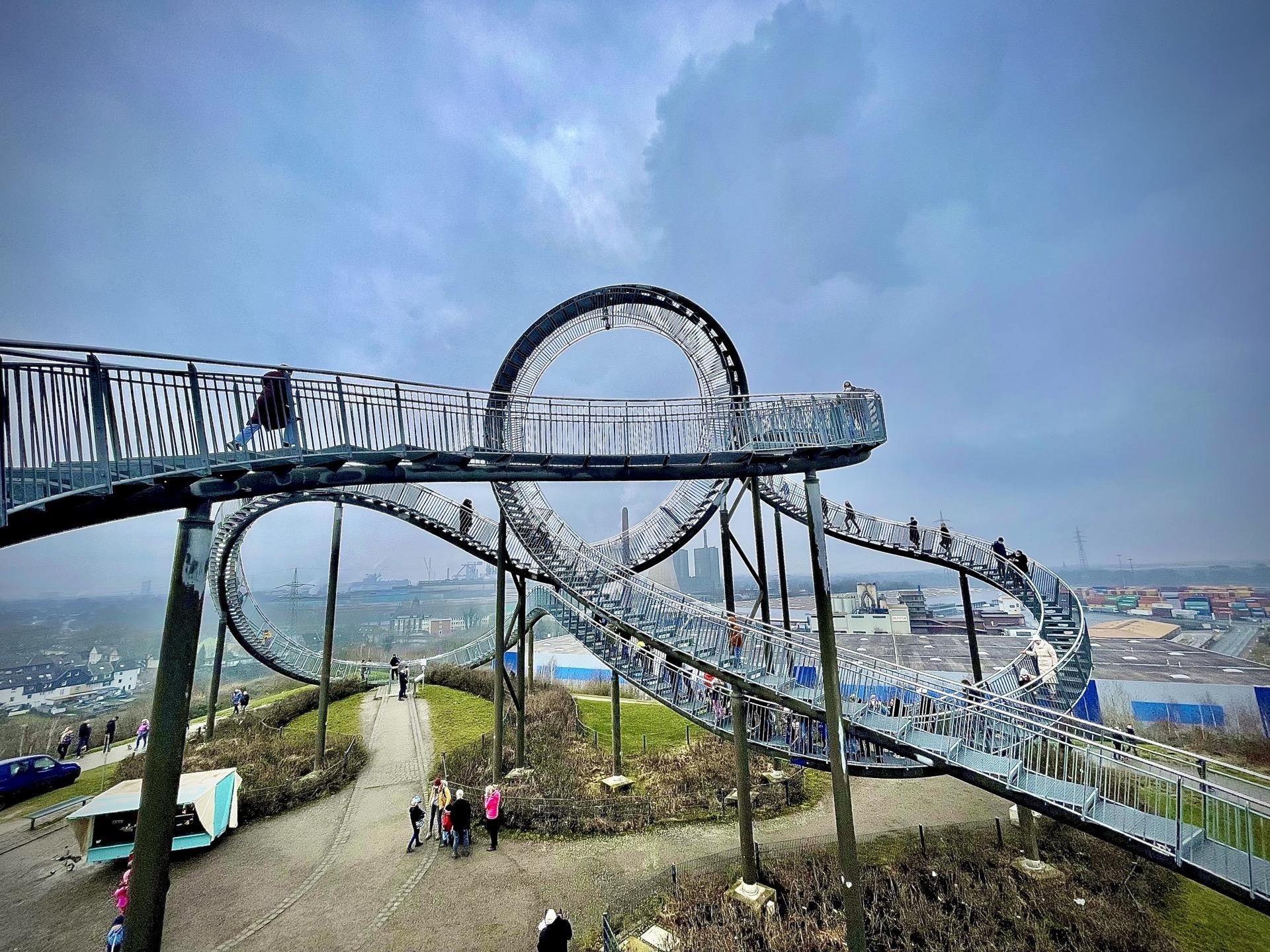 Pedestrian bridge with an intricate loop structure stands over a grassy hill with several people walking across and around it. A clouded sky serves as the backdrop, and the landscape extends into an industrial area with multiple containers in the distance.