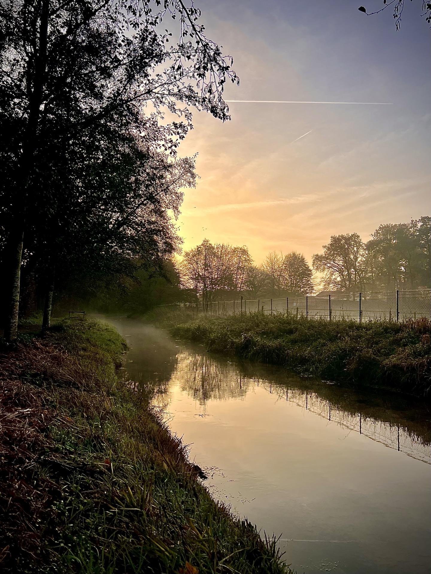 Sunrise over a serene landscape featuring a gentle stream flanked by grassy banks and lush trees. A thin layer of mist hovers above the water's surface, enhancing the scene's calm tranquility. The early light casts warm hues and softens the silhouettes of the trees against a sky streaked with the faint trail of an airplane. A metal fence lines one side of the stream, contrasting with the natural elements.