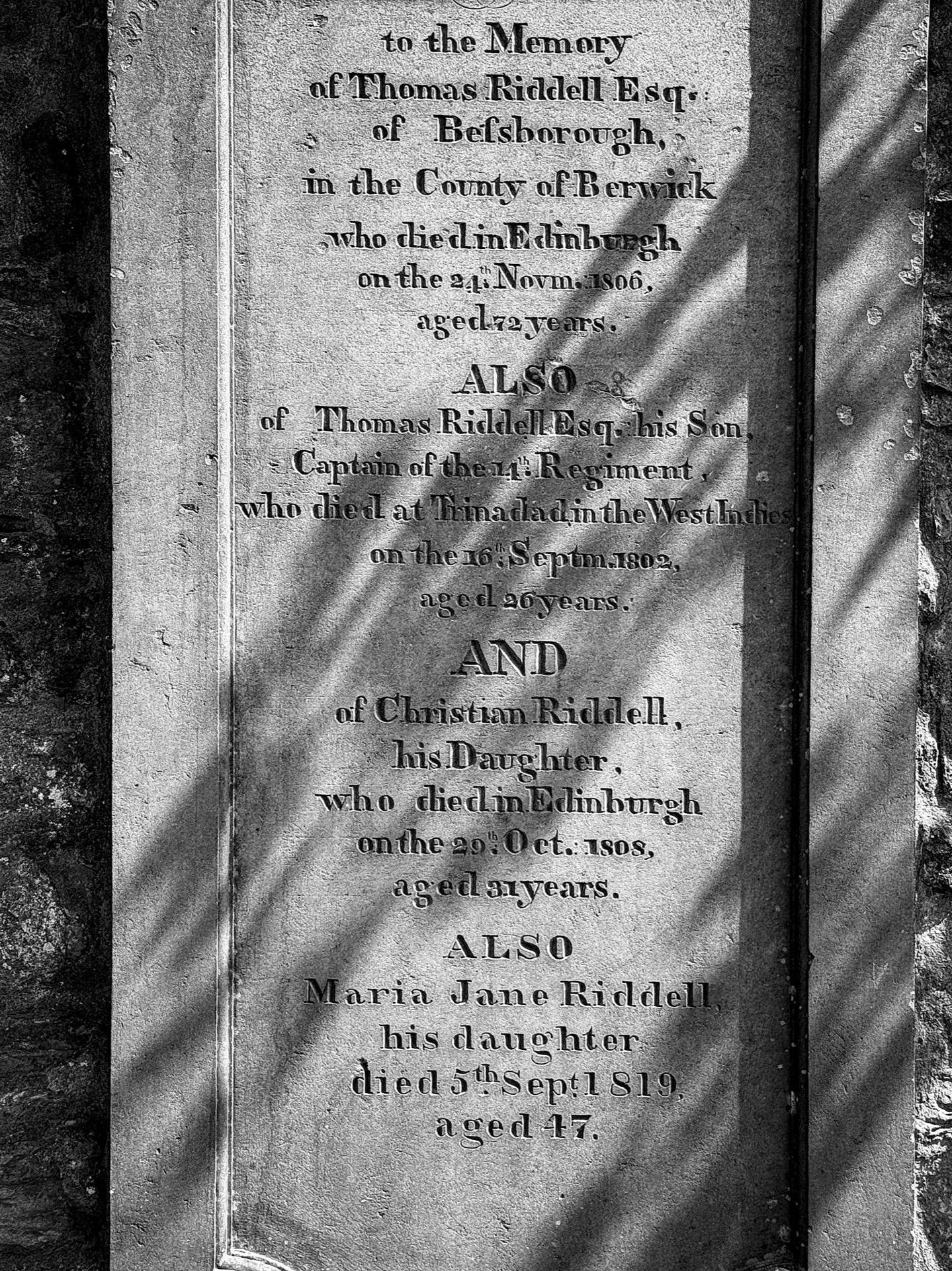 Close-up of an aged, weathered gravestone with engraved text memorializing Thomas Riddell Esq. and his family. The engraving lists names, dates, and ages at death, mentioning locations such as Edinburgh and Trinidad. Part of the stone features a shadow cast by an unseen object, emphasizing the texture and erosion on the stone's surface.
