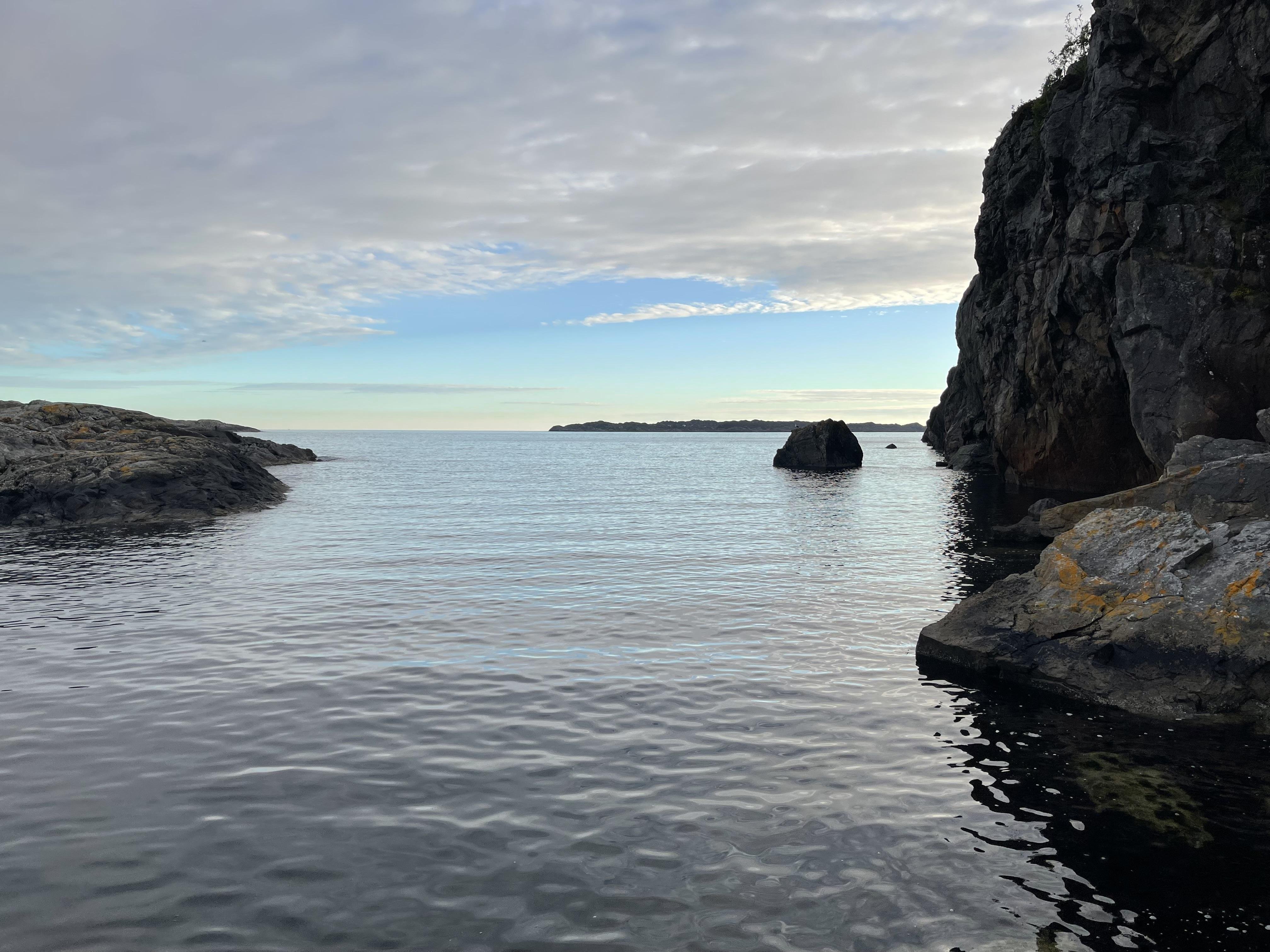 A tranquil seascape with a calm sea bordered by rugged cliffs on each side, leading into the open water. The sky is overcast with scattered clouds, casting a soft light over the scene. A small rocky islet is visible in the middle distance.