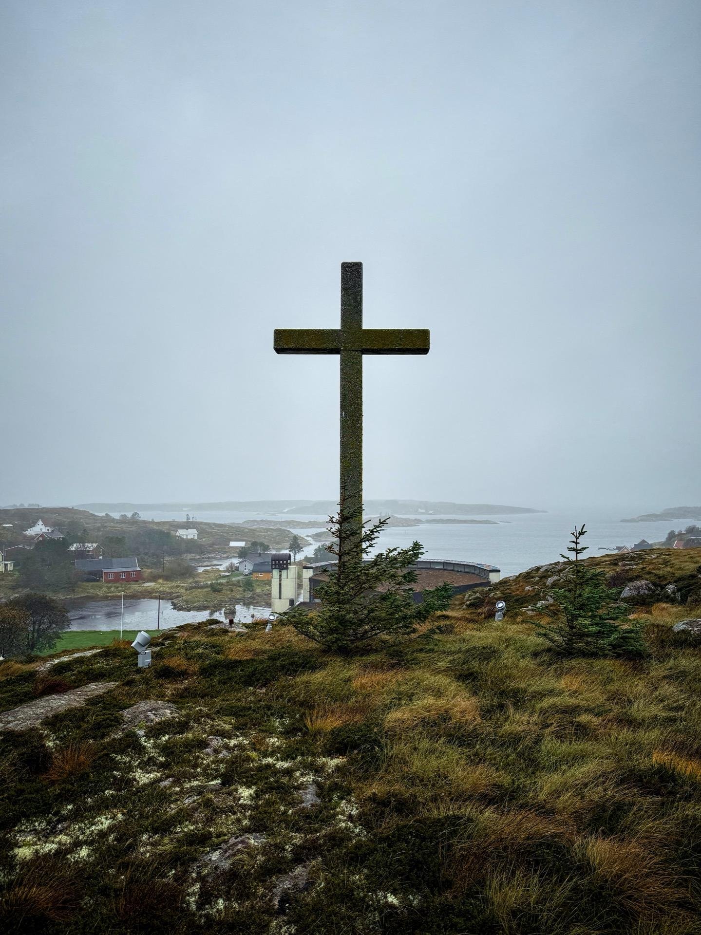 Large stone cross on a hilltop overlooking a coastal village with scattered buildings, under overcast skies. The foreground features rocky terrain with sparse vegetation and a few small trees, leading to a view of a calm bay surrounded by faint, distant landforms shrouded in mist.