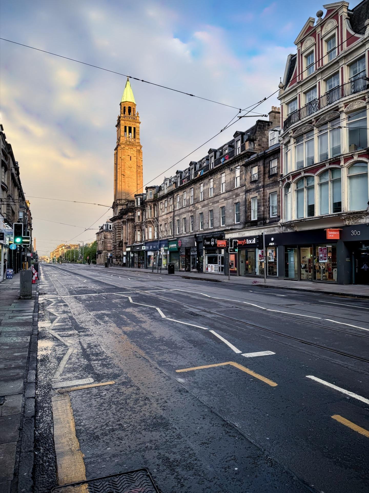 Early morning view of a deserted urban street lined with Victorian-style buildings and a prominent church tower rising in the background. The street features tram tracks and wet asphalt reflecting the soft, cloudy sky above.