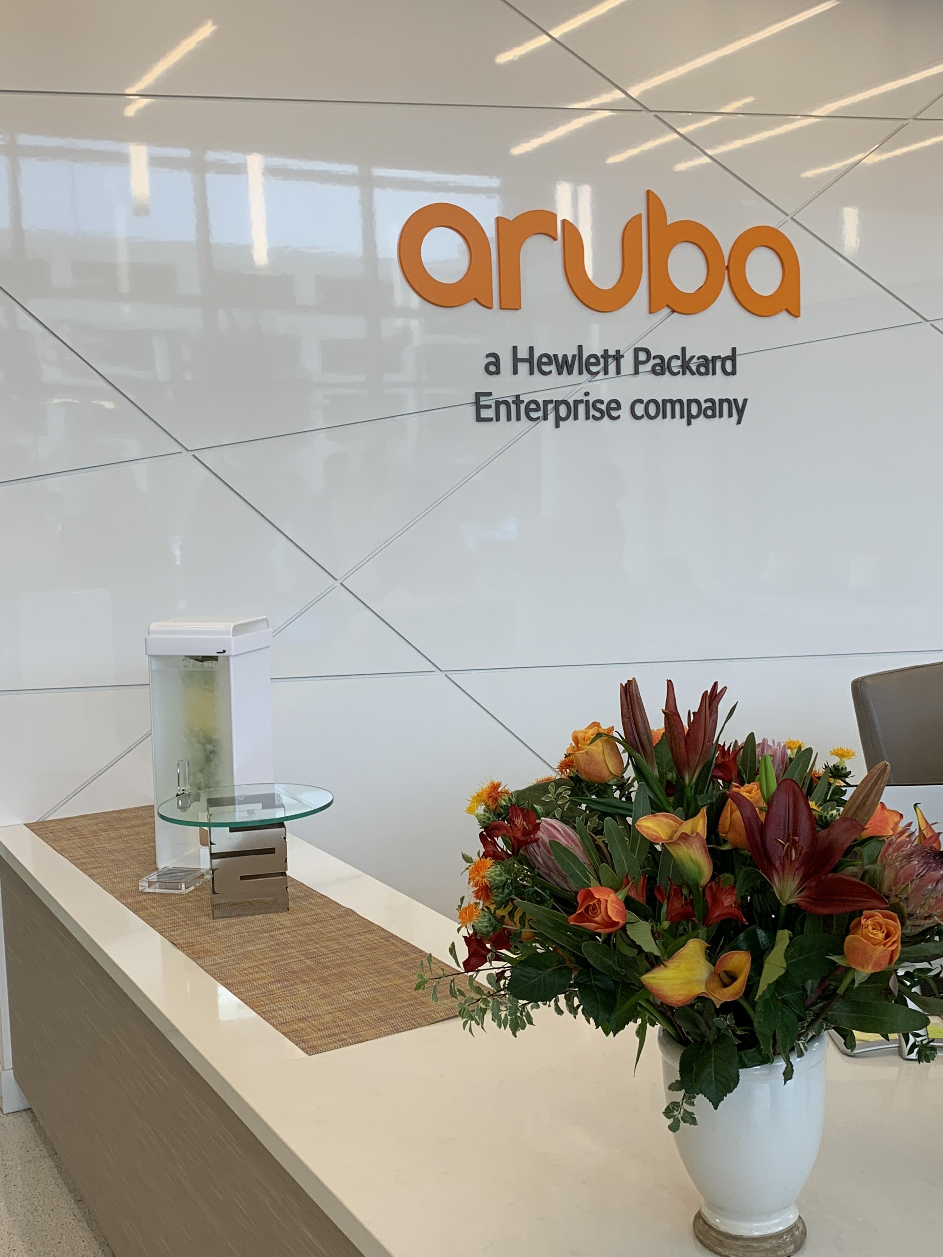 Orange "Aruba" logo with the subtitle "a Hewlett Packard Enterprise company" on a white wall above a reception desk. On the desk, there is a water dispenser with lemon slices and a large bouquet of flowers featuring roses and lilies in a white vase.