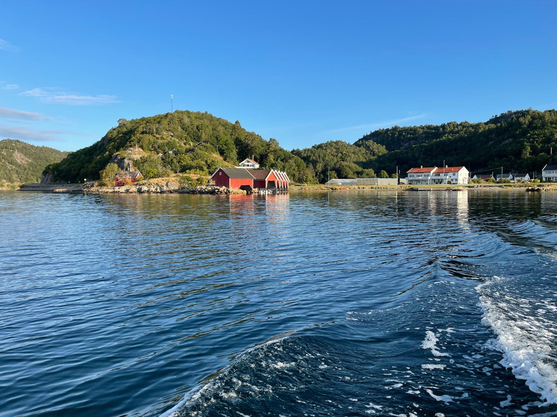 View of a calm sea with a wake from a boat, featuring a small island with a green forested hill and traditional red and white buildings on the coastline under a clear blue sky.