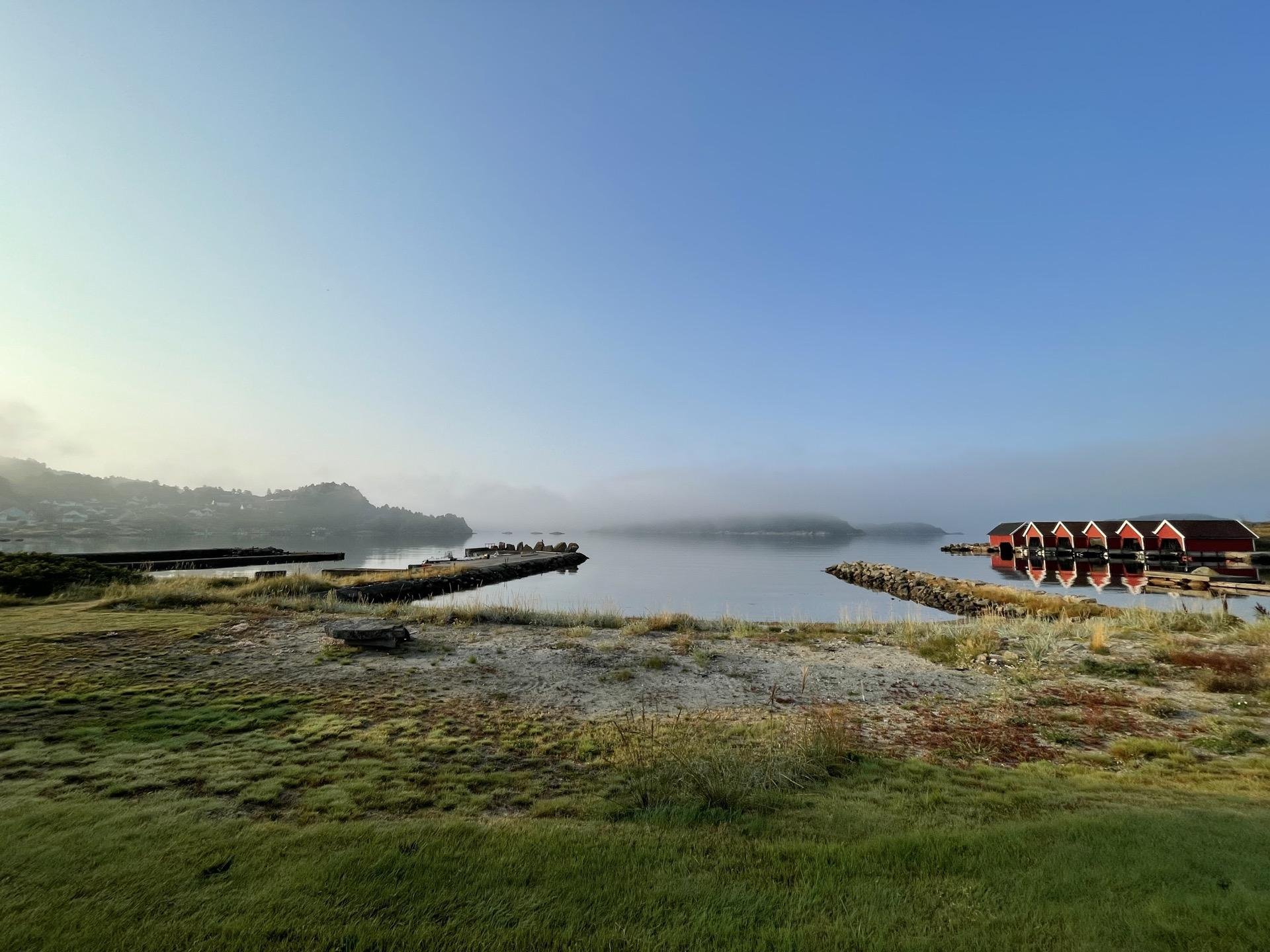 Early morning landscape featuring a calm lake partially obscured by mist, with a grassy shoreline in the foreground. A wooden pier extends into the lake on the left, and a row of red-roofed cabins on a rocky breakwater is visible on the right. The sky is clear and bright above.