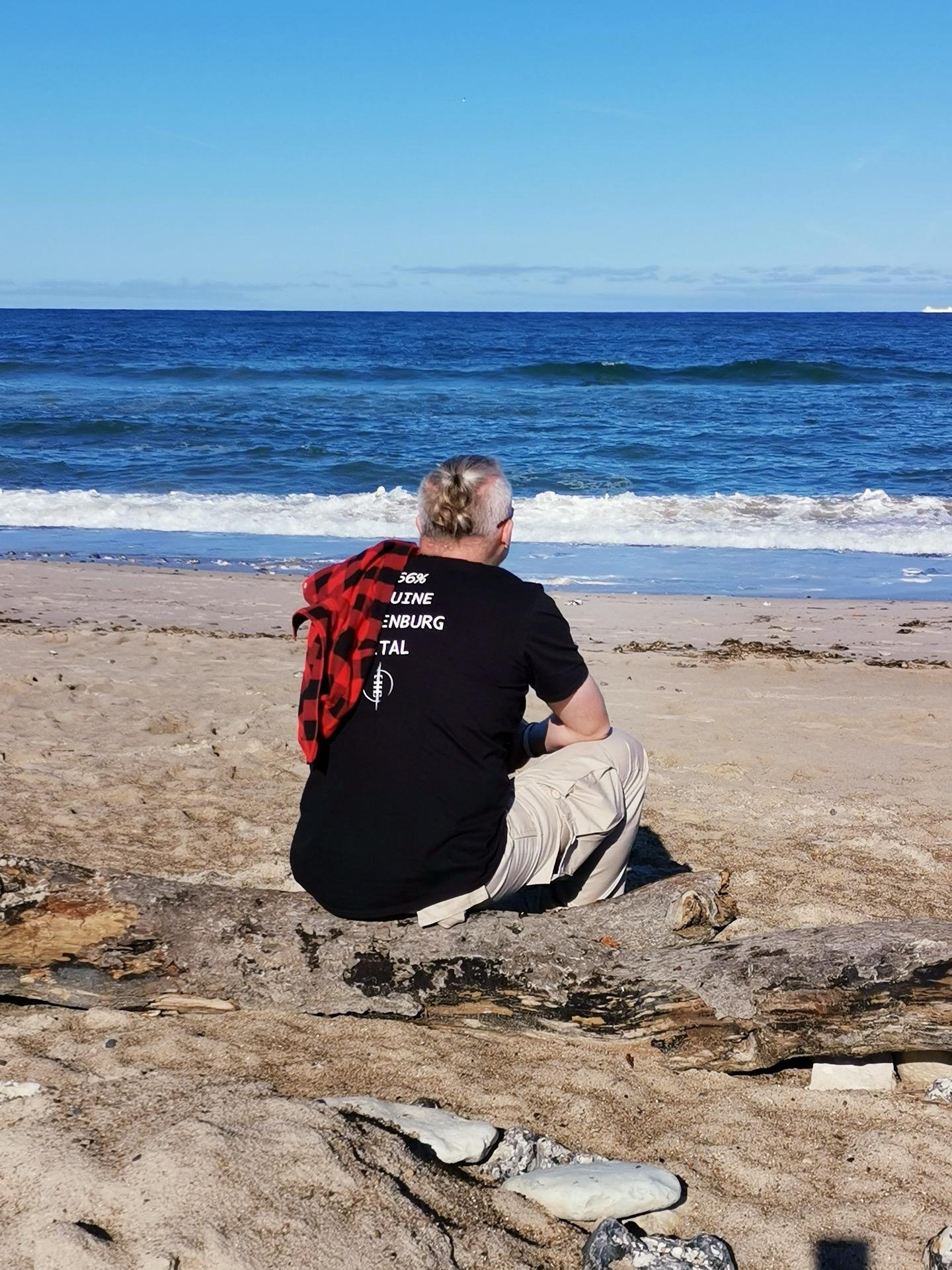 Man seated on a sandy beach facing the ocean, his back to the viewer. He wears a black t-shirt, beige pants, and has a red-checked shirt resting on his shoulder. A large driftwood log lies in the foreground with the blue sea and clear sky in the background.