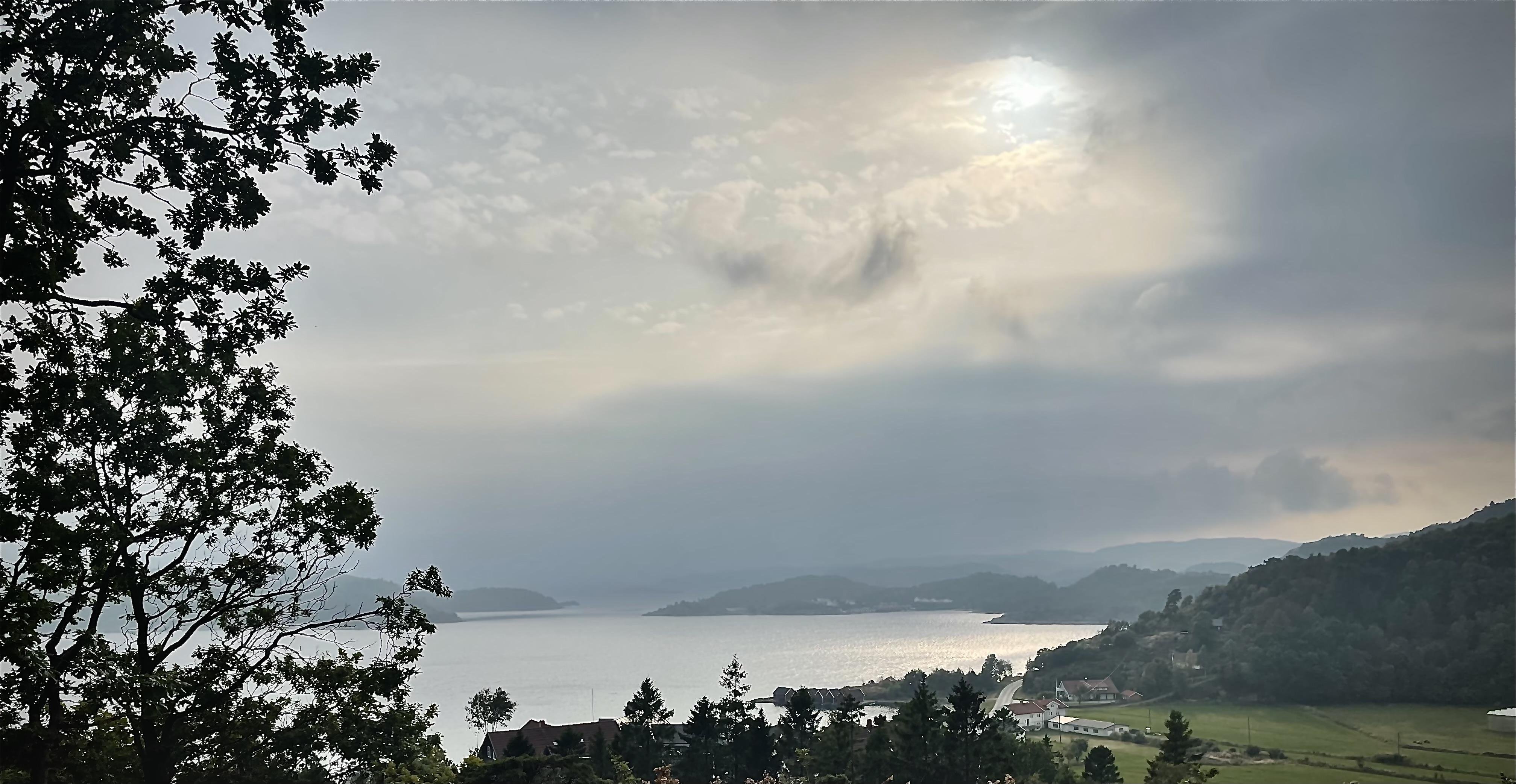 Overcast sky with the sun peeking through the clouds above a large lake surrounded by forested hills and a few visible buildings in a rural setting.