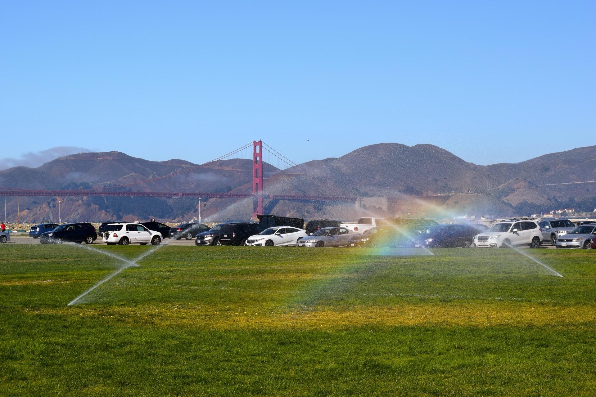Green grassy field with sprinklers creating a rainbow effect in the foreground. A parking lot full of cars extends towards the background, with the Golden Gate Bridge and mountains visible under a clear blue sky.