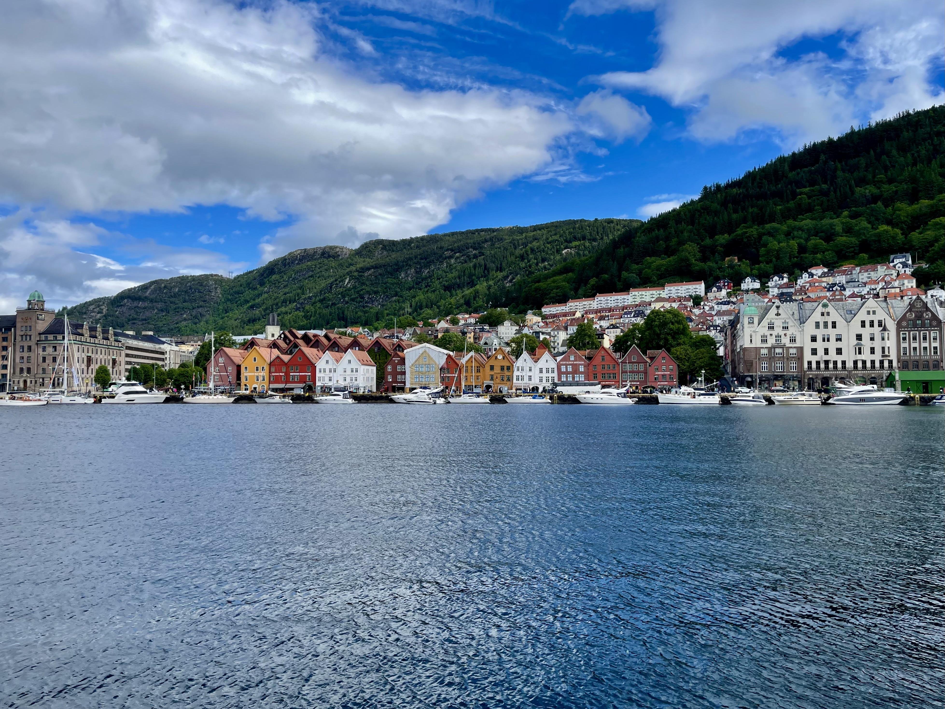 Colorful traditional buildings line a waterfront with boats moored in front under a cloudy sky, backed by a steep, tree-covered hillside.
