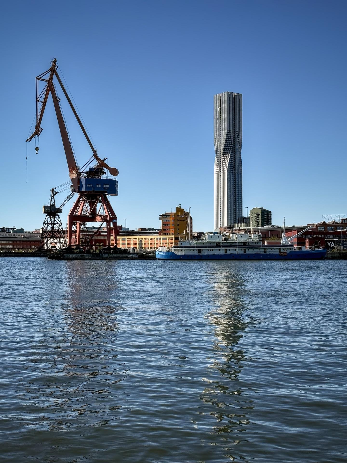 Large red crane on a dock to the left and a tall, twisted skyscraper in the background under a clear blue sky. A body of water in the foreground reflects subtle ripples and the buildings, with a blue boat moored at the harbor.