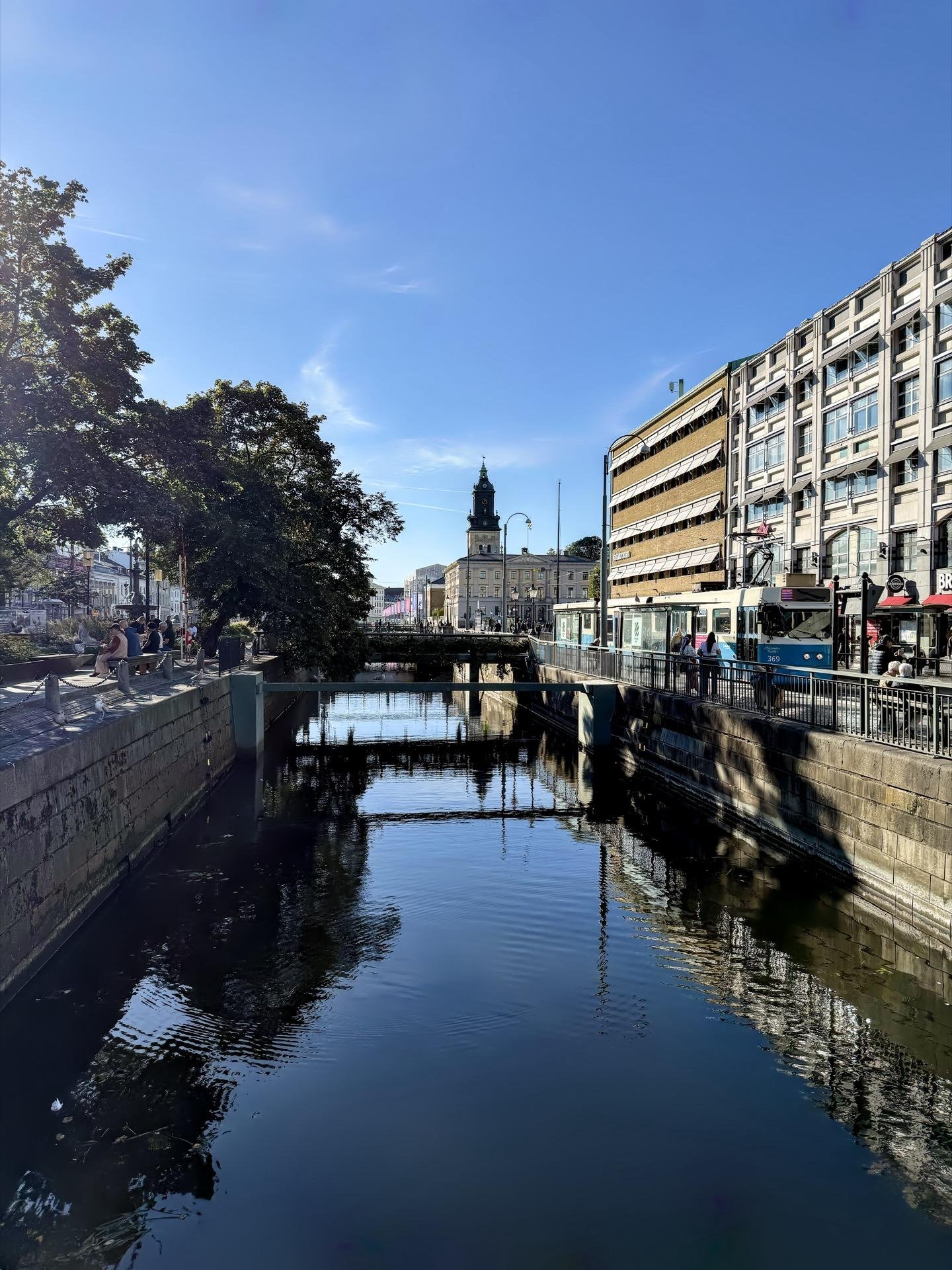 Urban canal flanked by modern buildings on the right and older, ornate architecture featuring a visible clock tower in the distance. The reflection of the sky and structures is visible in the water. A clear blue sky and scattered trees add natural elements to the scene.