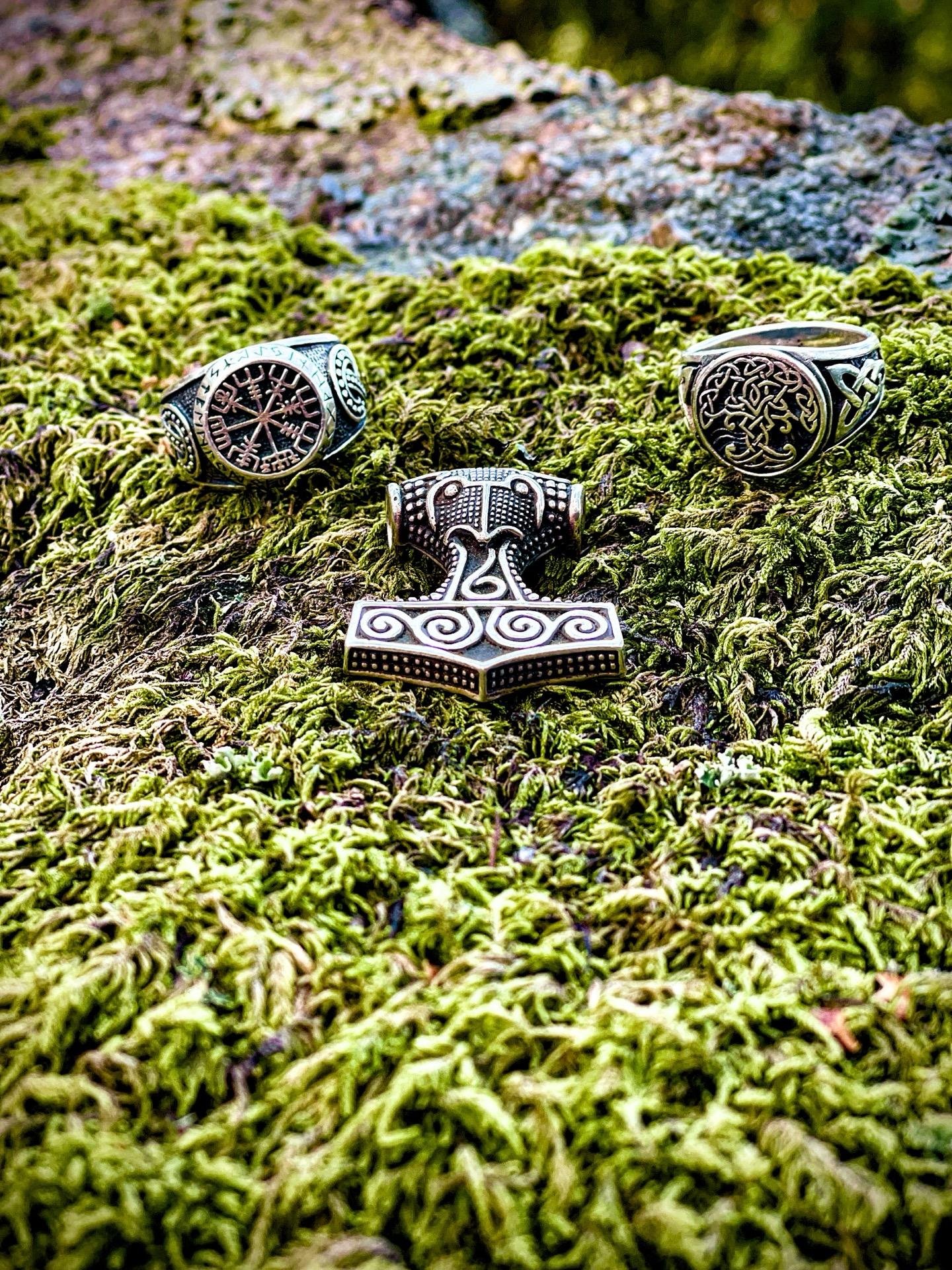 Three ornate, Viking-style rings and a pendant rest on a bed of lush green moss, with each piece featuring intricate Nordic designs. The moss texture is dense and vivid, providing a natural contrast to the metallic sheen of the jewelry.