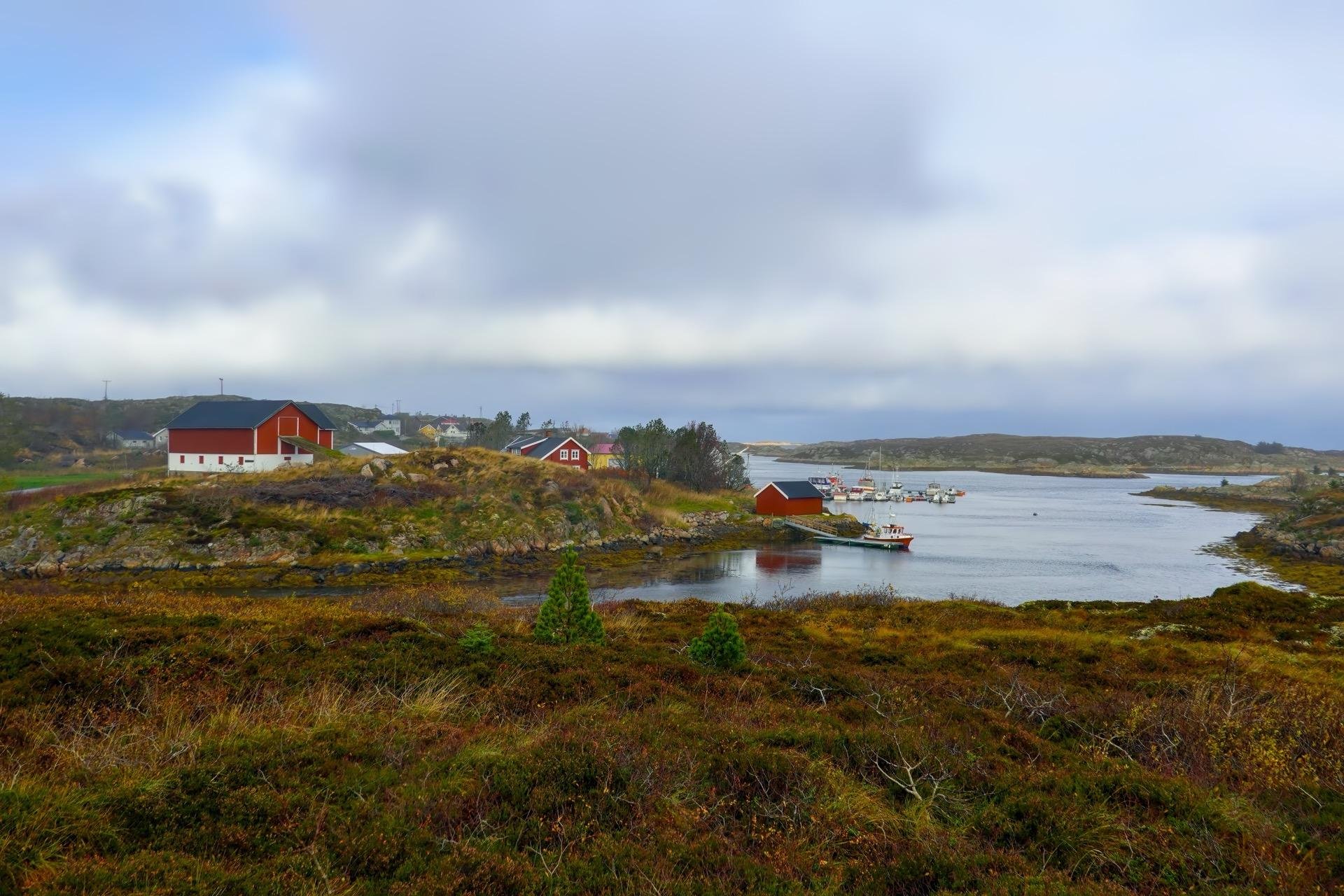 Overcast sky above a rocky coastal landscape with a cluster of buildings. A vivid red house stands prominent among smaller red and white buildings, interspersed with patches of grass and low vegetation. A serene water body in the foreground reflects a boat and the cloudy sky, surrounded by sparse greenery.