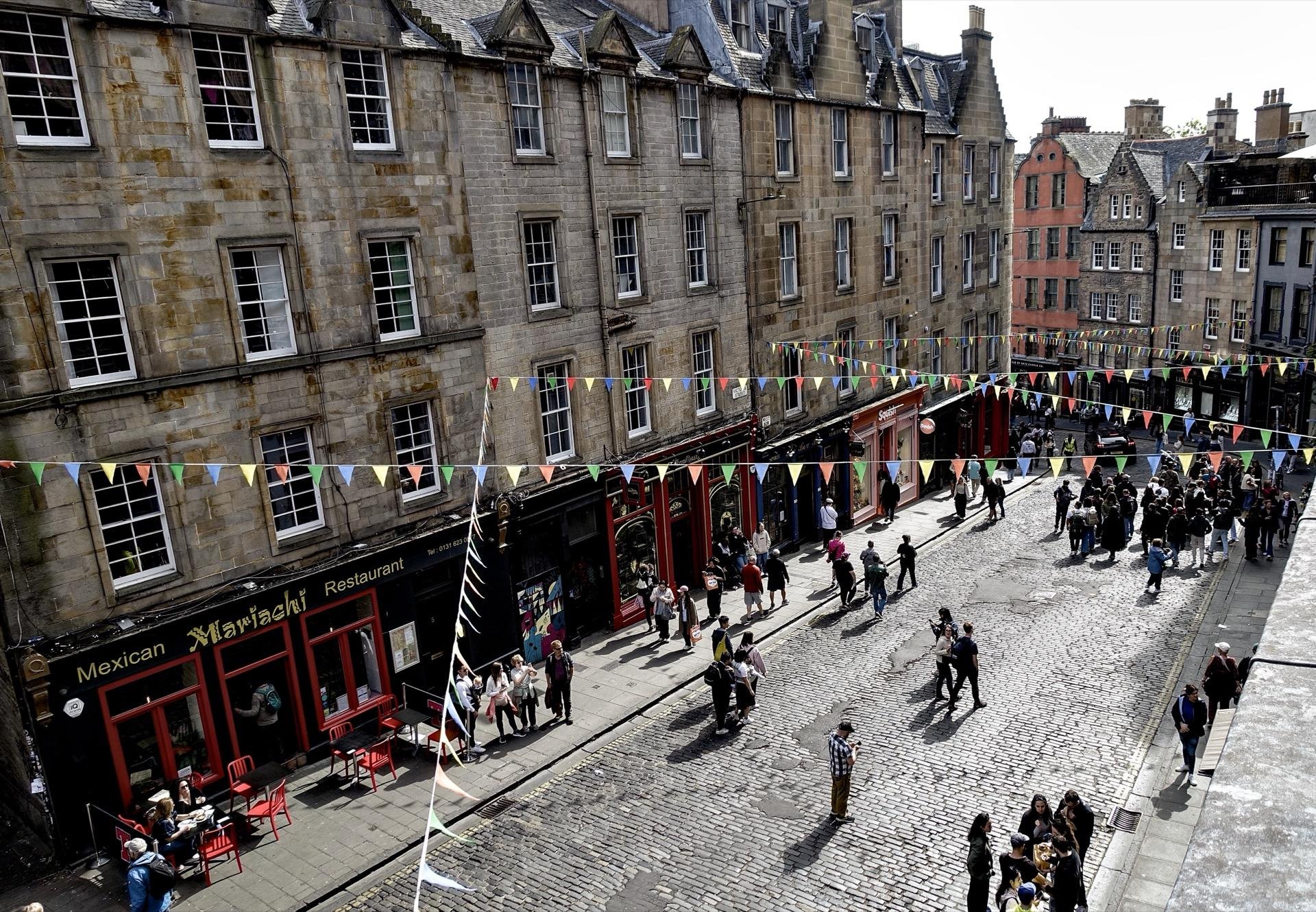 Split-view of two cobblestone streets lively with pedestrians in a historic European city. The left image features a row of aged stone buildings with ground floor businesses, including a Mexican restaurant with red outdoor seating under colorful flag bunting. The right image captures a wider street scene bustling with people, lined with various shops under a series of multicolored flags stretched across from building to building, and a notable red and orange building visible in the background.