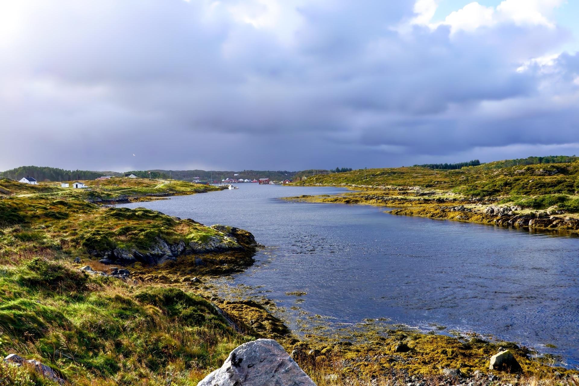 Coastal landscape featuring rocky terrain and sparsely vegetated patches surrounding a calm body of water, under a cloudy sky. Distant houses visible, possibly a small village or settlement.