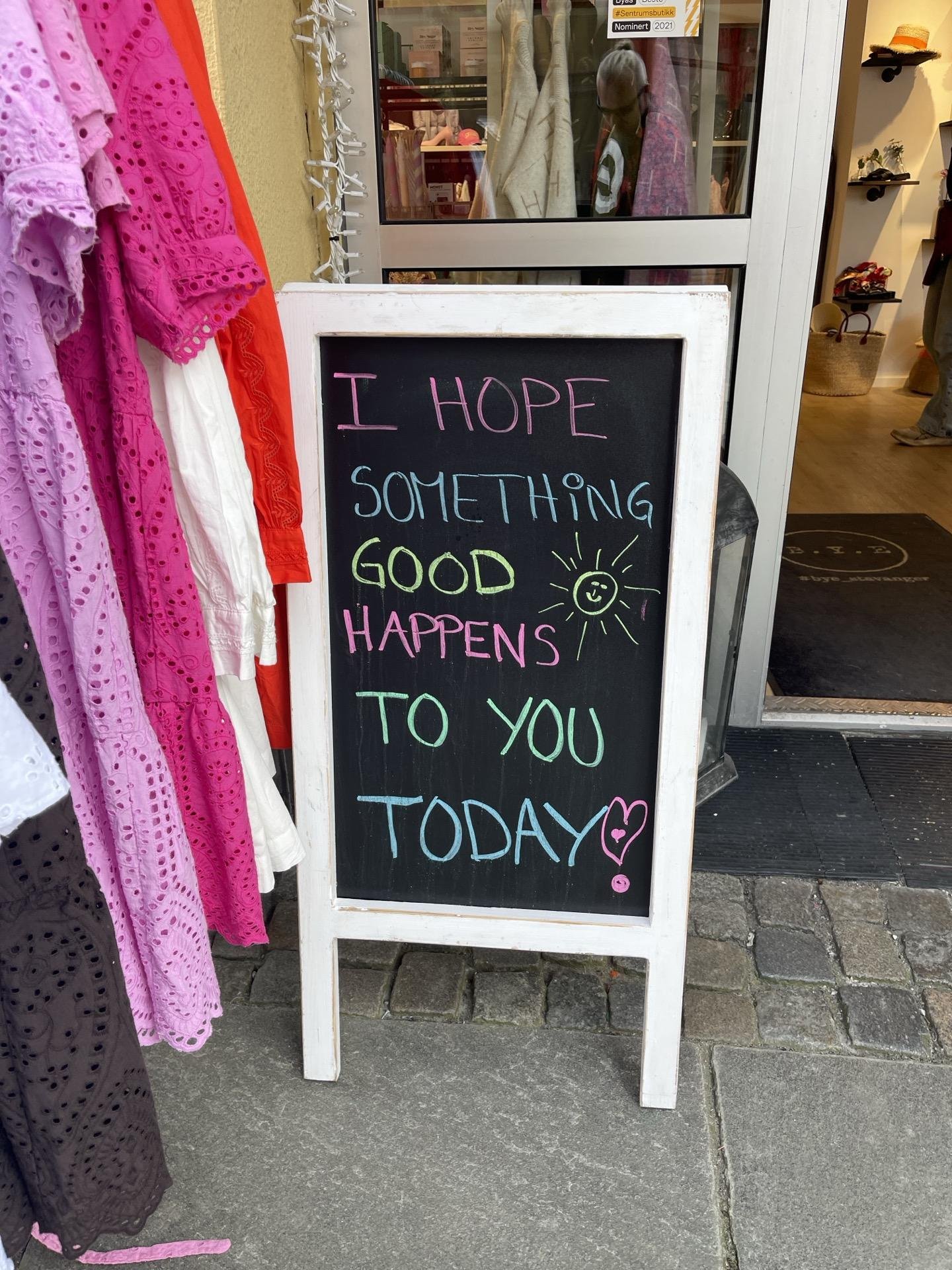 A sidewalk signboard with a chalk message stating "I HOPE SOMETHING GOOD HAPPENS TO YOU TODAY" in mixed colorful lettering. The sign is positioned outside a store, partially flanked by brightly colored clothing, including a pink and a purple garment on the left. The background shows the entrance of the store with visible garments and reflections of the bustling street.