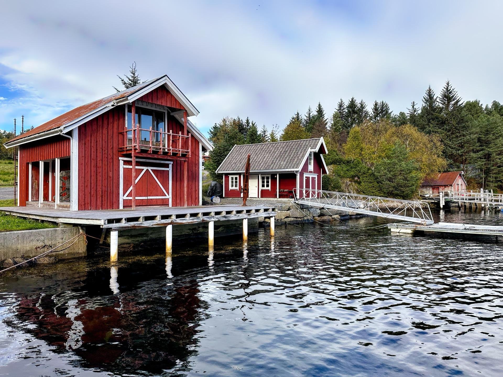 Two red wooden buildings beside a calm body of water, one built on stilts directly over the water with a balcony facing a deck, and the other on land connected via a small bridge. Both buildings are surrounded by green trees under a cloudy sky.