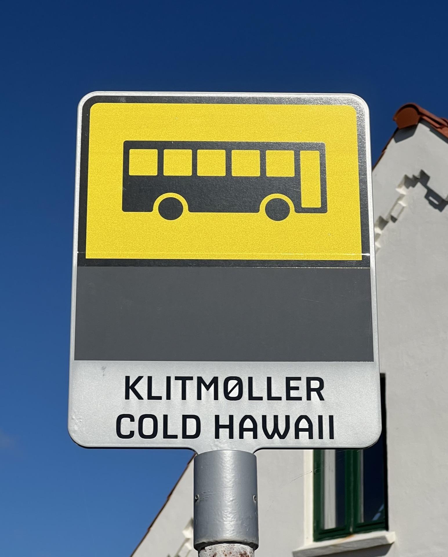Bus stop sign with a bright yellow background and a black silhouette of a bus, mounted on a pole. Below the bus image, a grey panel reads 'KLITMØLLER COLD HAWAII' in black capital letters. A clear blue sky and part of a white building with a red roof are visible in the background.