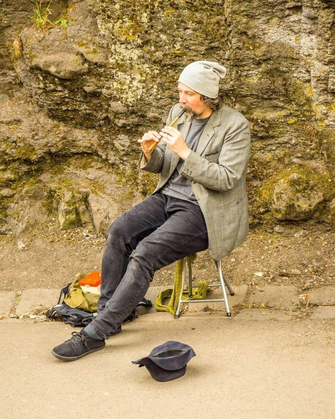 A man with a beard, wearing a gray knitted hat and a tweed jacket, sits on a metal folding chair against a rocky background while playing a wooden flute. He wears black pants and casual shoes, and a dark cap is placed upside down on the ground in front of him, likely for collecting tips. Beside him, a backpack and a yellow cloth are visible on the pavement.