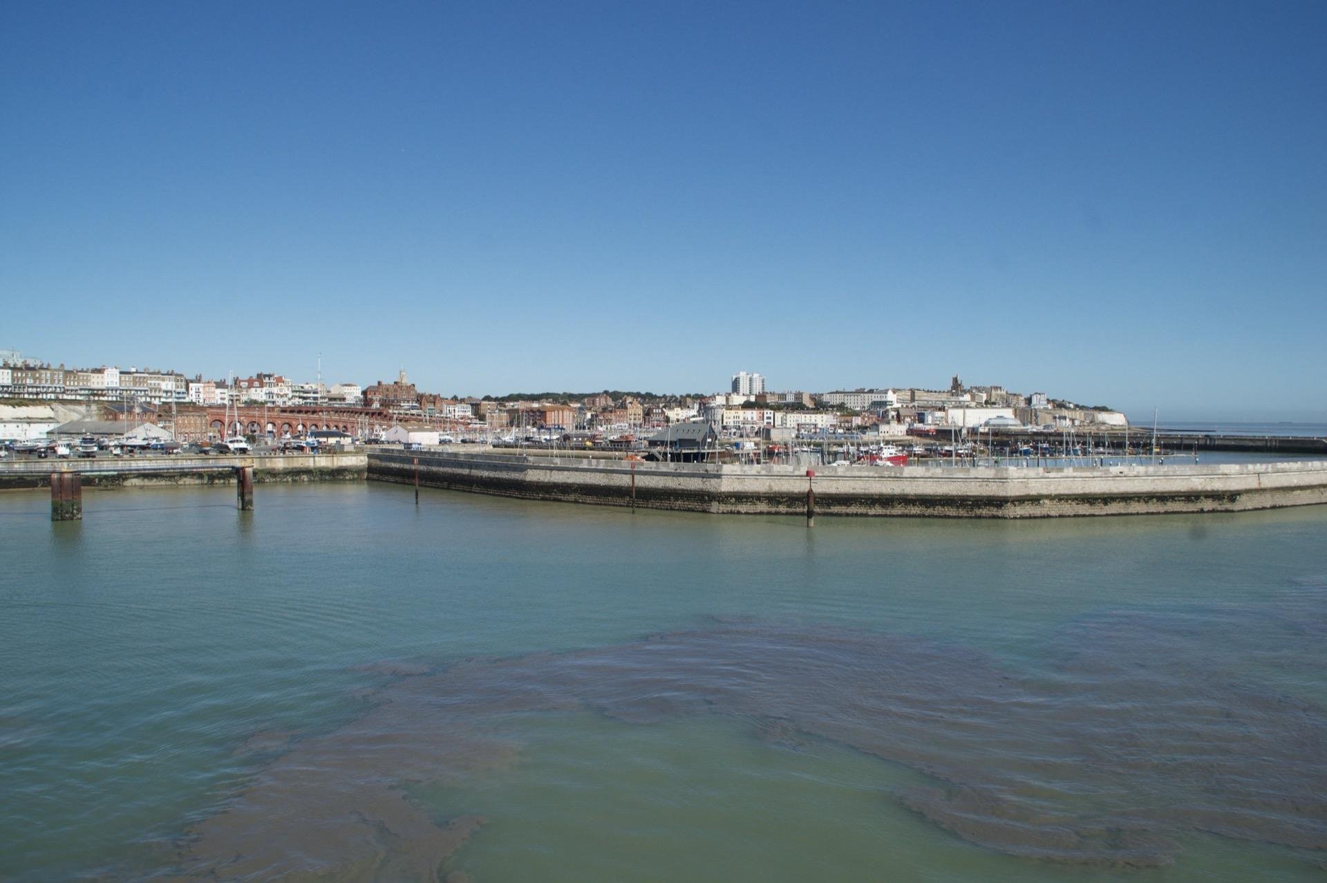 Wide angle view of a coastal town with clear blue skies over calm sea waters; the town features densely packed traditional buildings and modern structures visible behind a harbor filled with boats and bordered by a protective seawall.