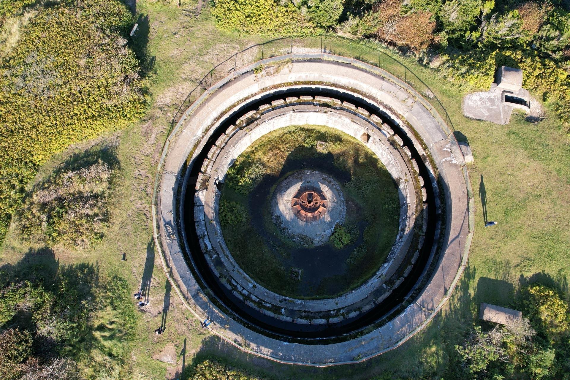 Aerial view of a circular abandoned structure surrounded by greenery. The structure features multiple concentric layers, each descending deeper into the earth with the innermost part containing rusty machinery. Patches of shrubs and trees border the structure and a few walkways lead out from it.