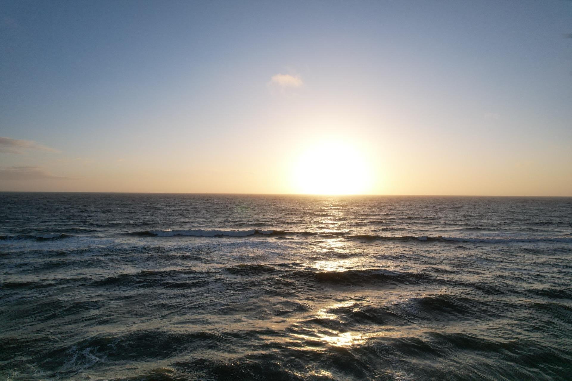Sunset over the ocean with the sun low on the horizon, casting a golden reflection on the rippling water. The clear sky above transitions from a soft yellow near the sun to a deeper blue at the top.