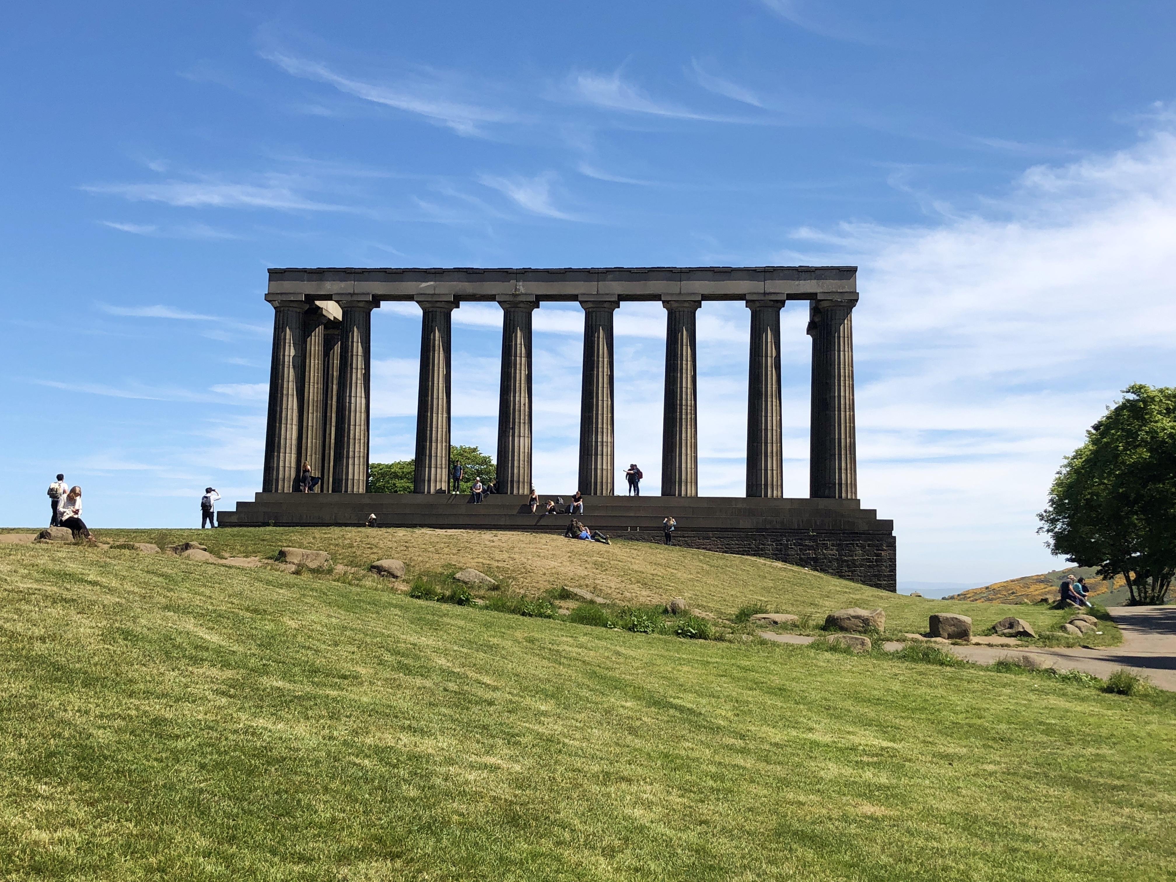 Large, Classical-style stone colonnade on a hilltop with several columns, under a blue sky. People are scattered around the site, walking on paths and sitting on the grassy slopes surrounding the colonnade.