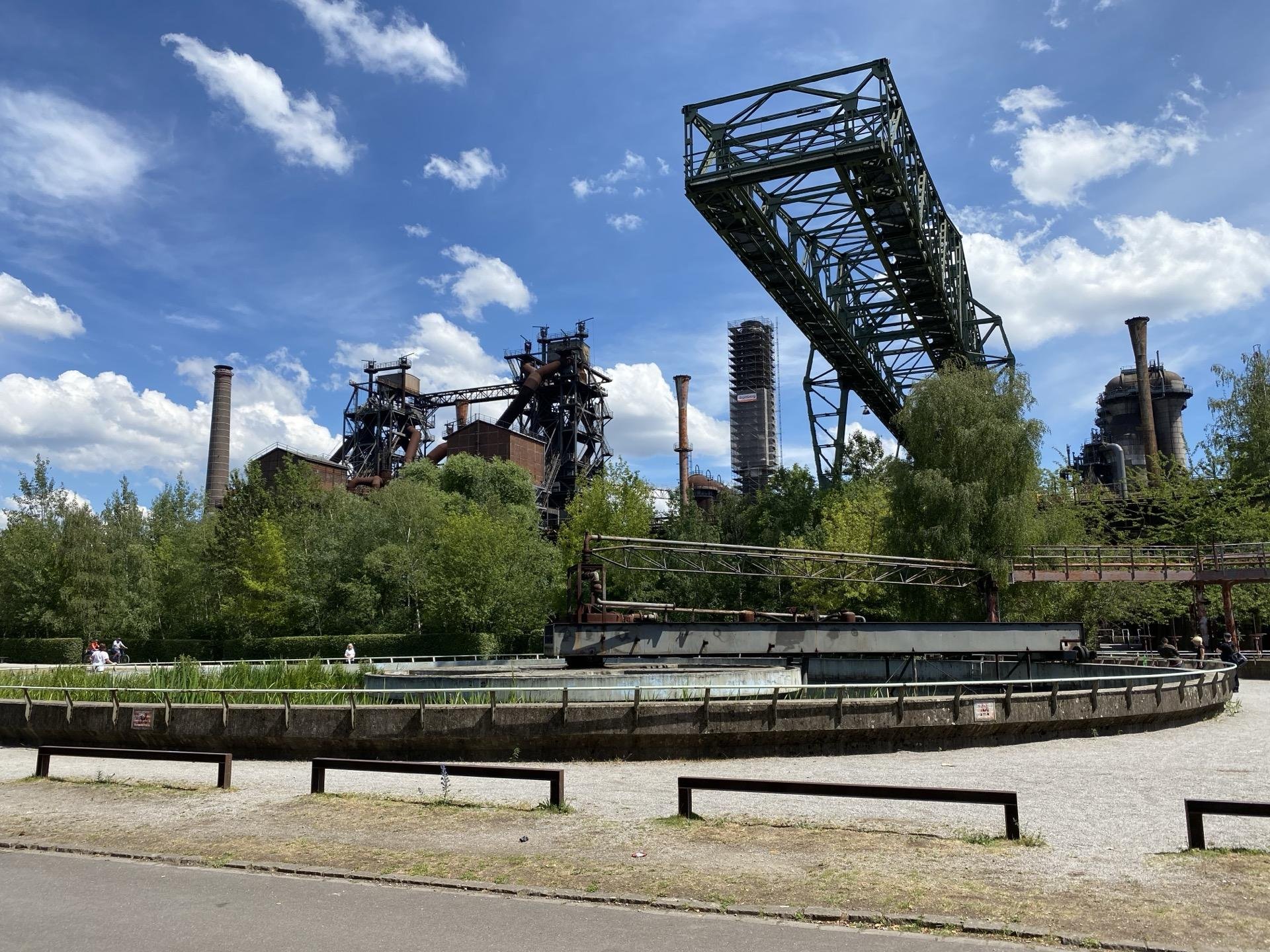 Old industrial site featuring large decommissioned blast furnaces and chimneys, with a prominent green metal bridge on the right. In the foreground, there is a circular pool and walking paths, surrounded by lush green trees under a partly cloudy sky. Several people are visible enjoying the park-like setting.
