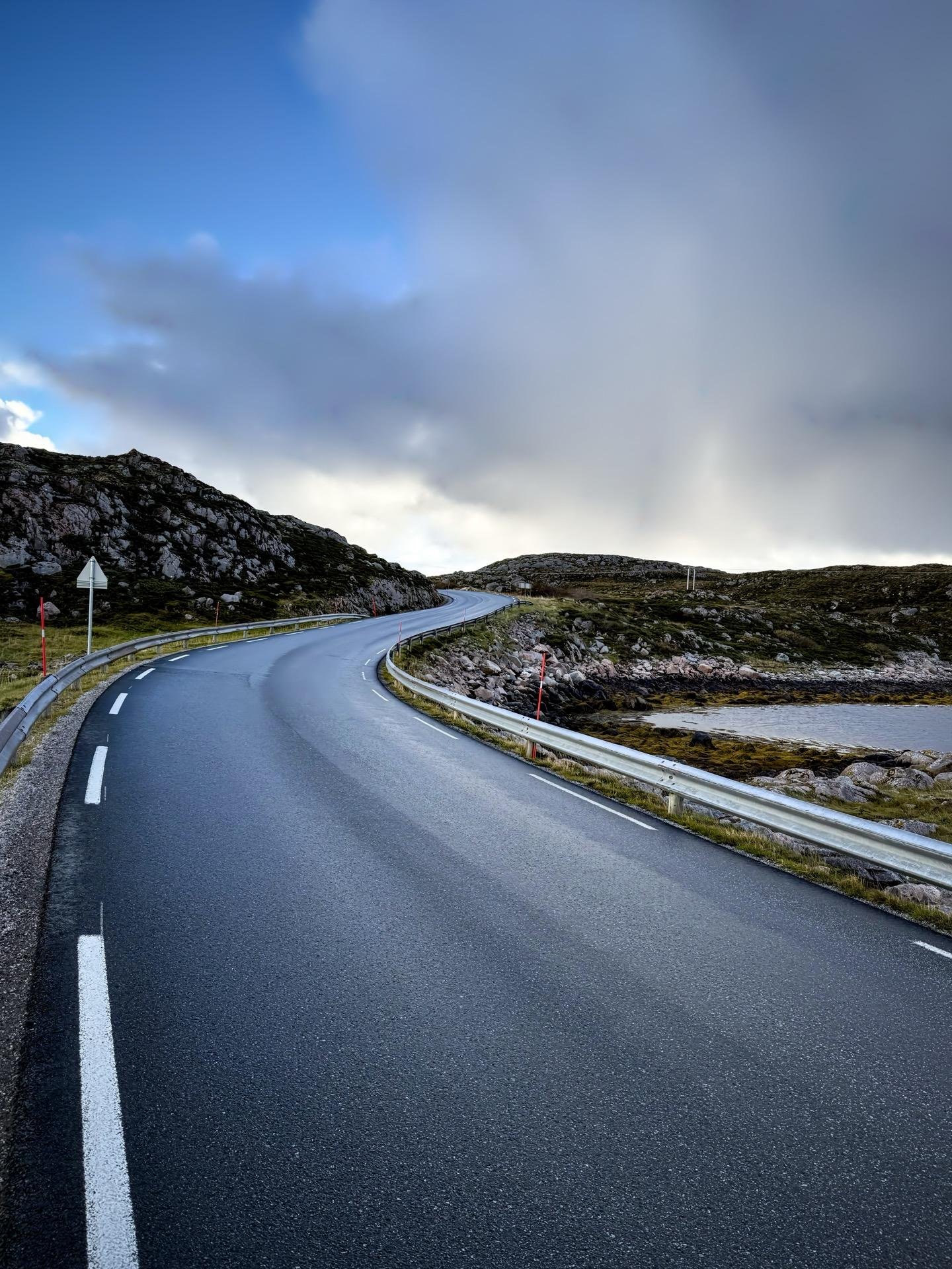 Curved road leading through a rocky landscape under a cloudy sky. White lane markings are visible on the asphalt surface, with red and white roadside barriers along the left side. The terrain is sparse with low grass and scattered rocks, and a body of water is partially visible beside the road.