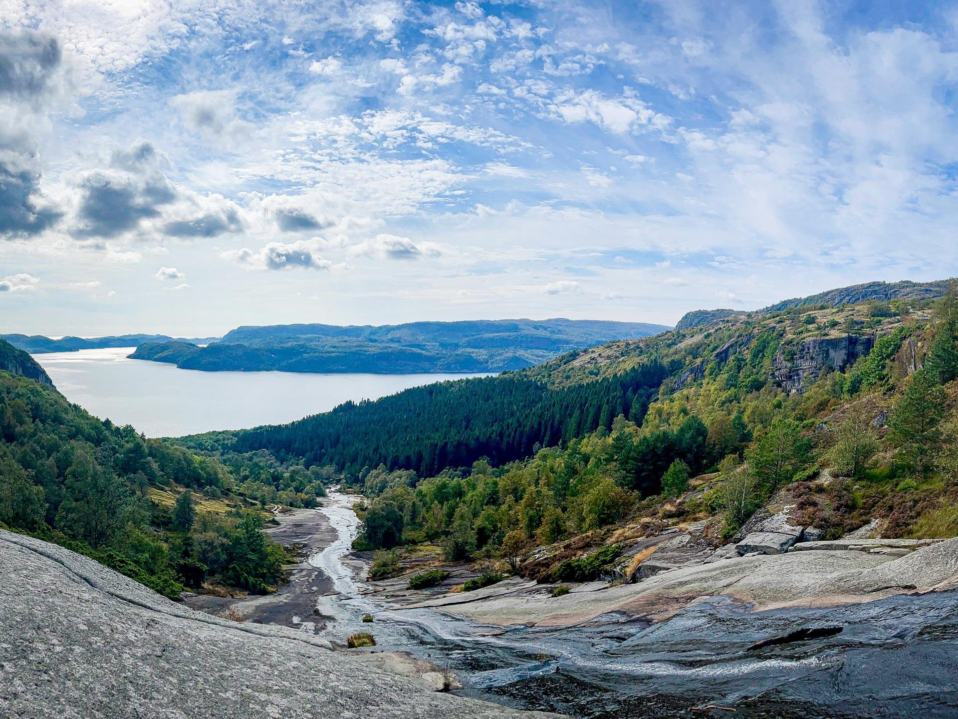 Panoramic view of a scenic landscape featuring a river winding through a forested valley, flanked by massive rock formations and hills, leading to a large body of water under a partly cloudy sky.