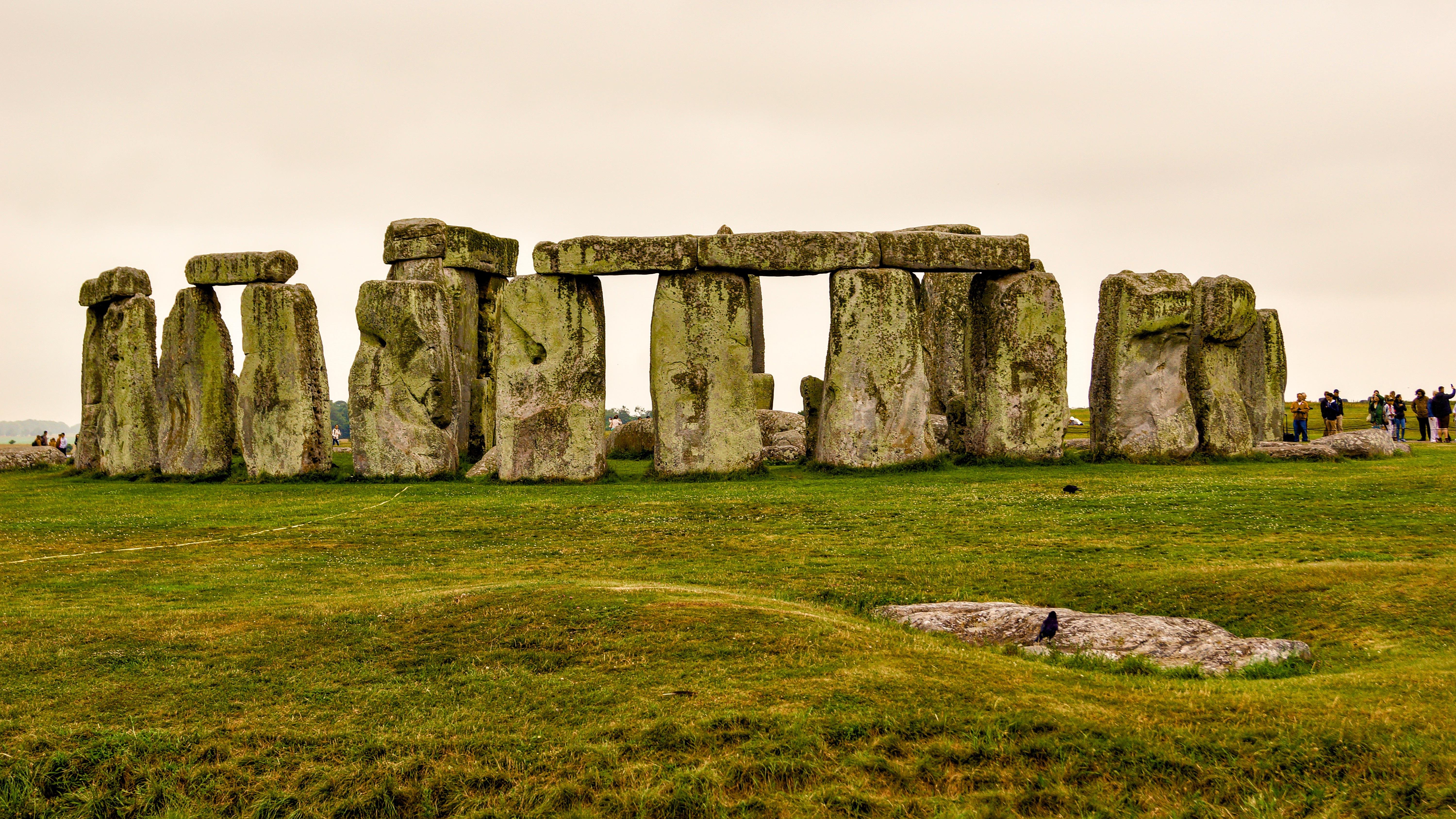 Stonehenge under overcast skies, featuring large standing stones arranged in a circular formation on a grassy field. Several people are visible in the background observing the monument.