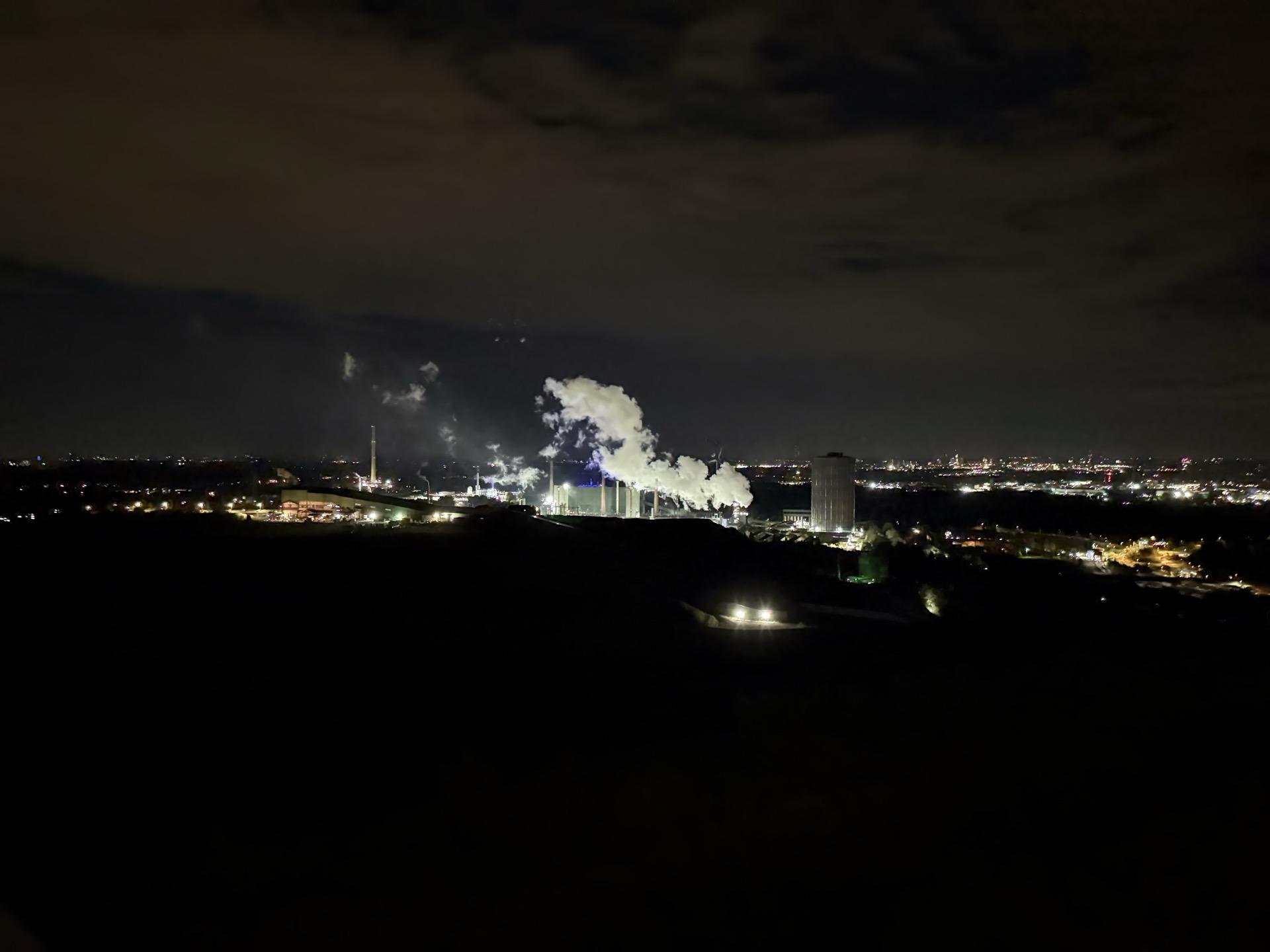 Nighttime view of an industrial area with multiple smokestacks emitting white smoke. Silhouetted hills in the foreground contrast with the brightly lit facilities and distant city lights. Cloudy skies hover above.