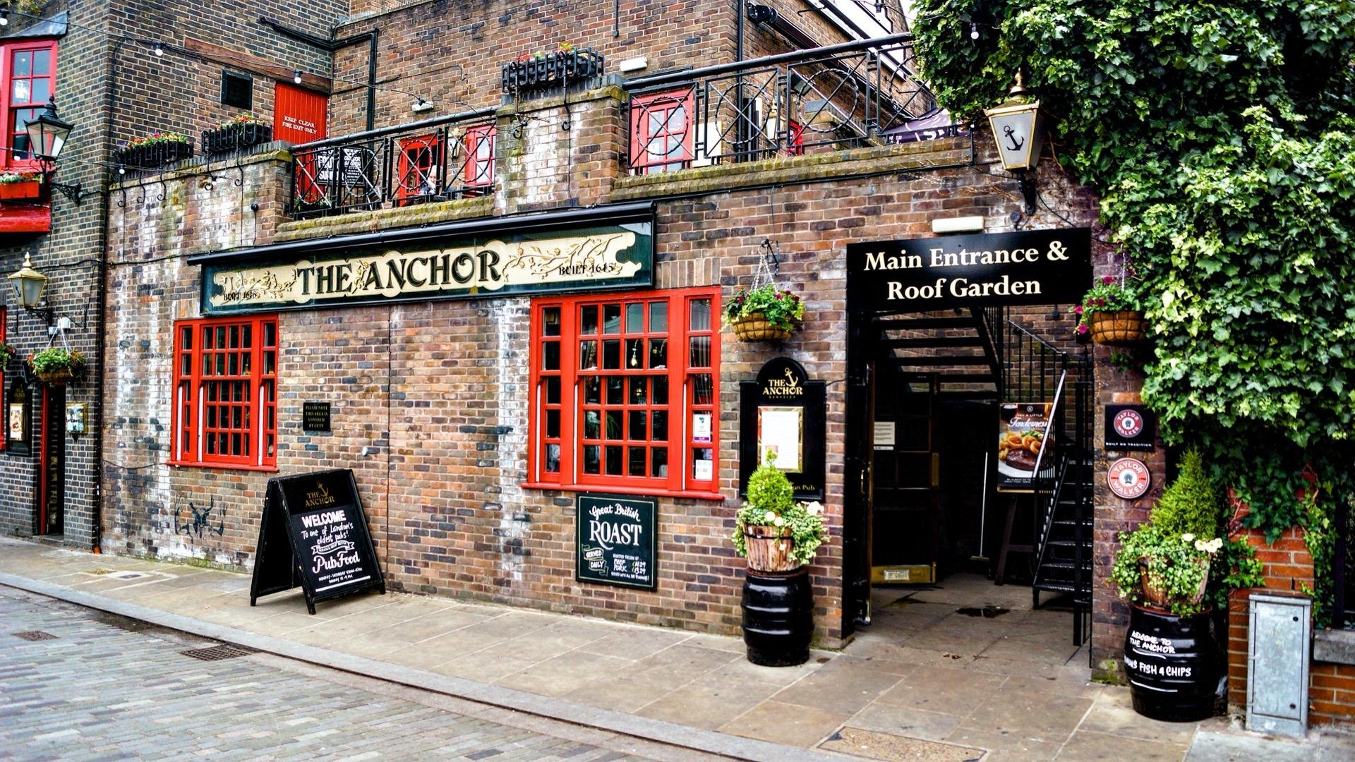 Brick facade of 'The Anchor' pub with red window frames and signage displaying the name and beverage offerings. A chalkboard sign advertises a Sunday roast near the entryway. The pub features intricate details like potted plants and vines climbing around and above the entrance, which is labeled "Main Entrance & Roof Garden." The street is cobblestoned, enhancing the traditional appeal.