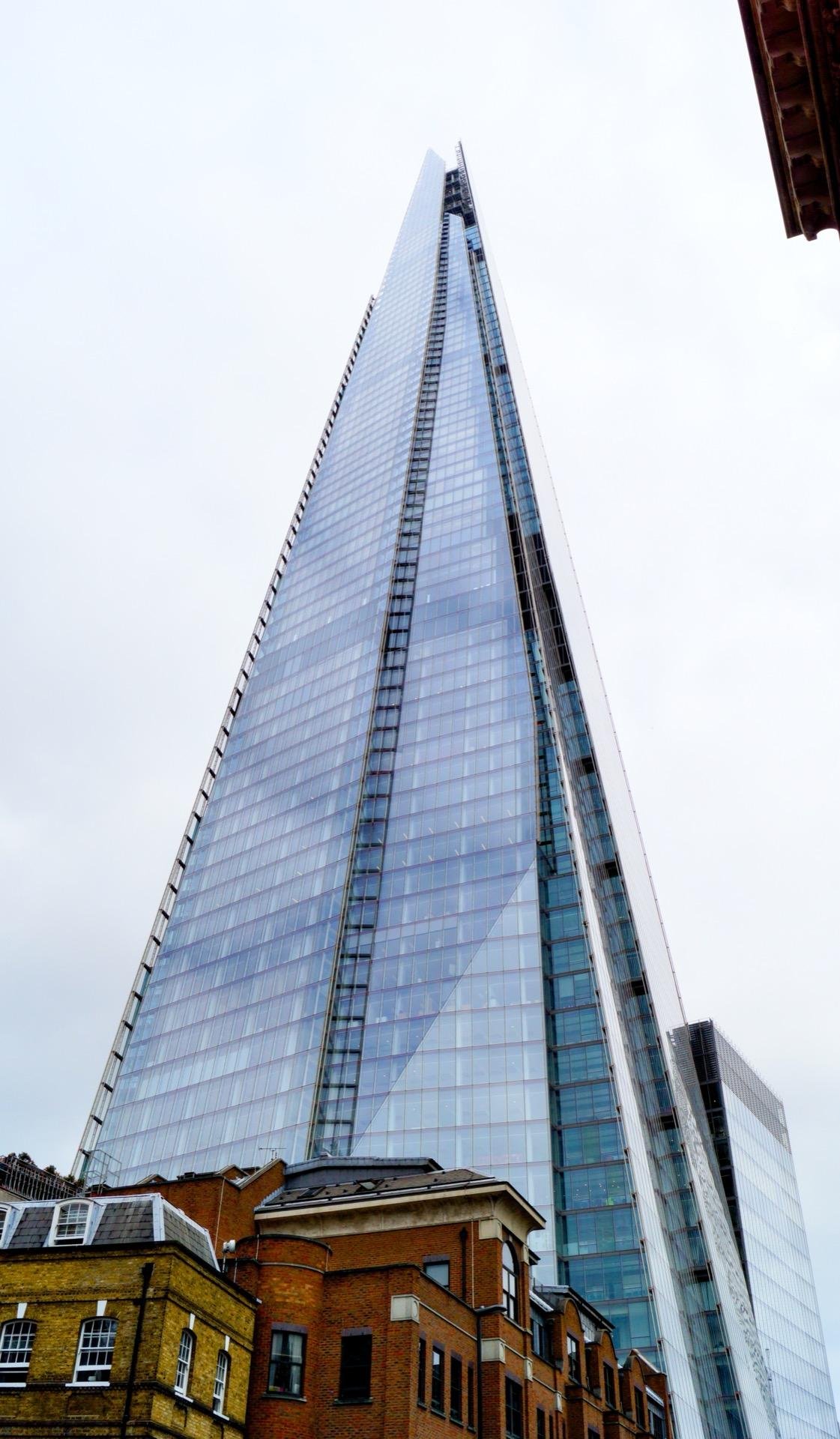 Tall glass skyscraper extending sharply into a cloudy sky, towering over traditional brick buildings in the foreground.
