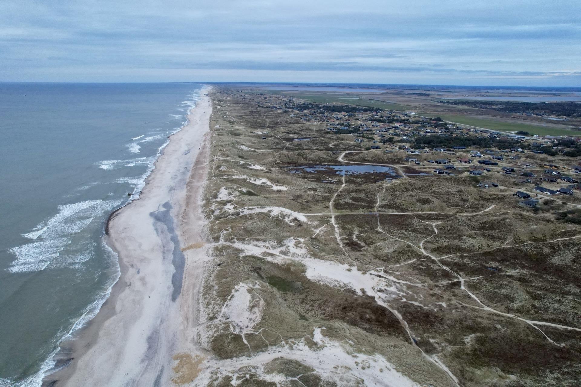 Aerial view of a narrow coastal barrier island featuring a sandy beach along the ocean side with intermittent waves, bordered by sand dunes and sparse vegetation. A cluster of buildings is present near the landward side, alongside patches of grassland and small roads, with a flat hinterland extending into the distance.