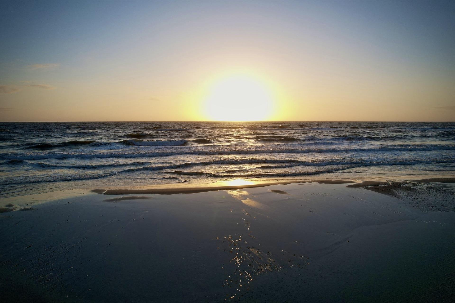 Sunset over a beach with the sun low on the horizon, casting a bright reflection on the wet sand and gentle waves rolling in from the sea.