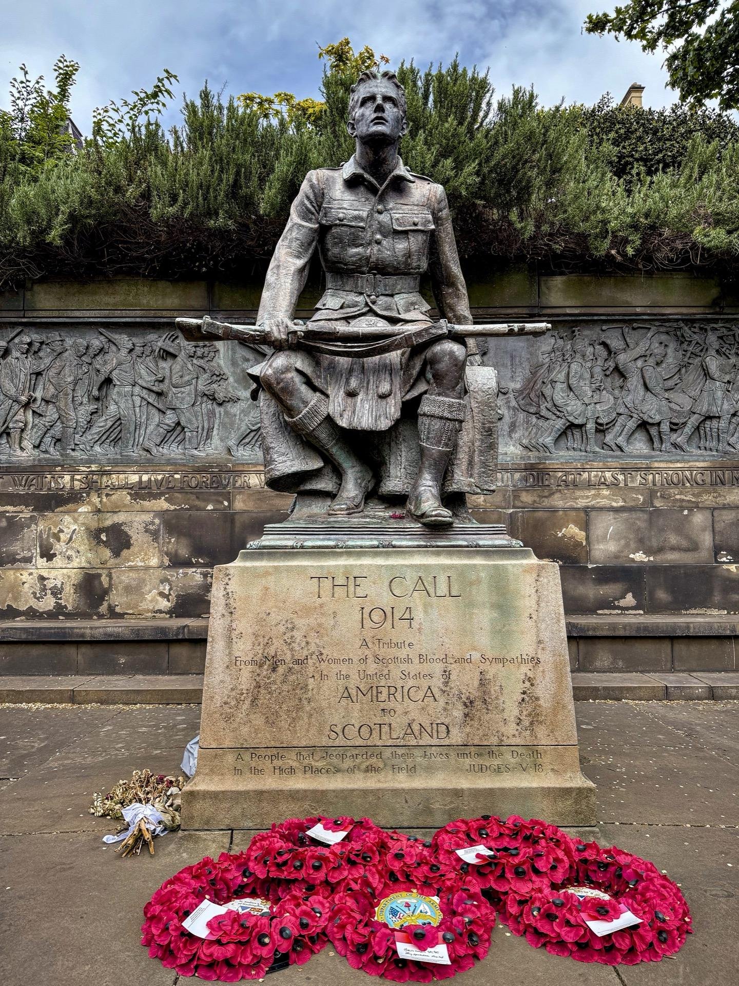Bronze statue of a kilted soldier seated on a stone base, holding a rifle, with a contemplative expression looking upwards. The base has an inscription reading "THE CALL 1914 - A Tribute From Men and Women of Scottish Blood and Sympathies in the United States of America to Scotland." Below the statue are red commemorative poppy wreaths with a few small bouquets. Behind the statue, a detailed bronze relief shows a historical battle scene. The setting is partially surrounded by green bushes under a gray sky.