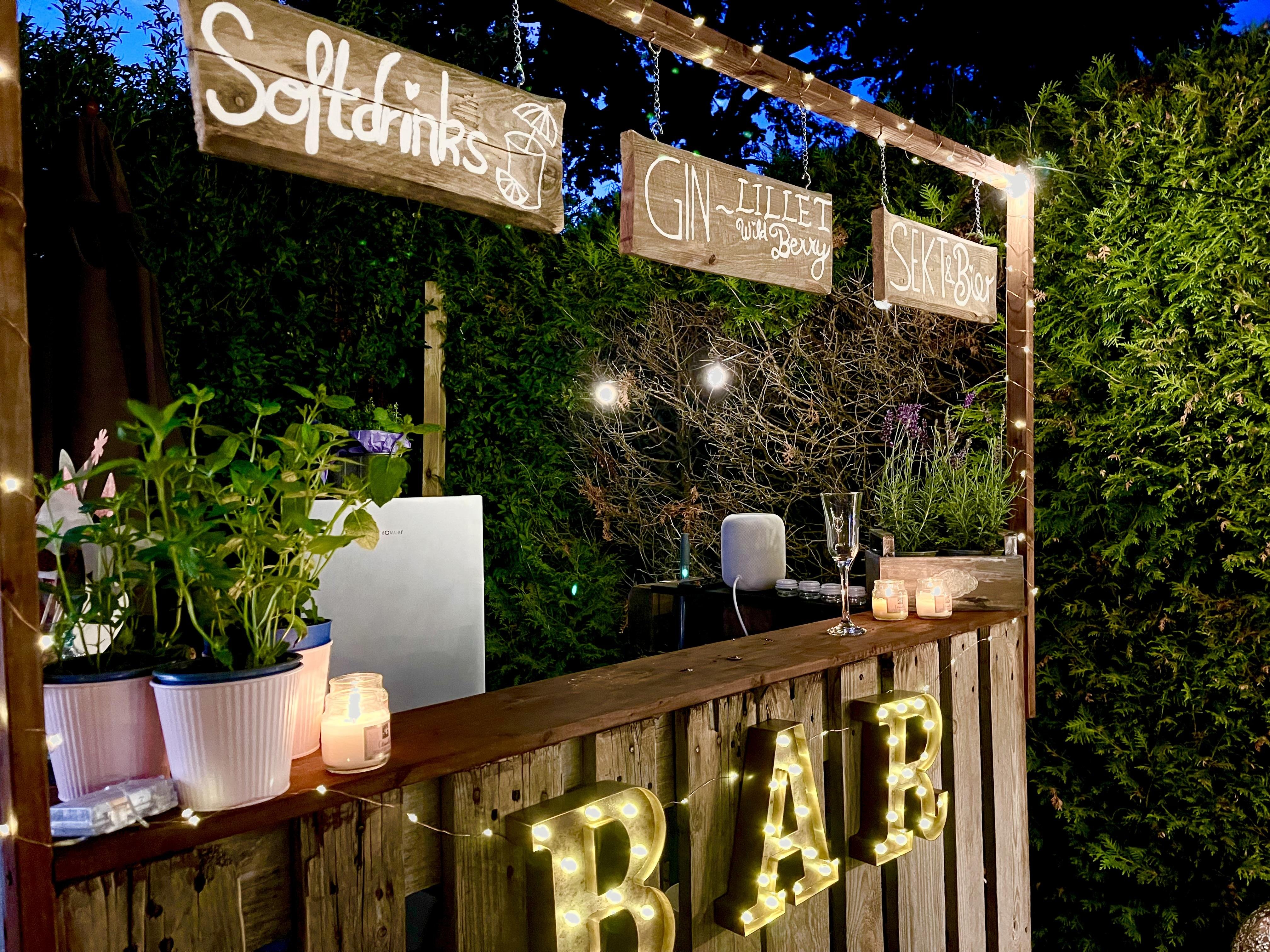 Outdoor bar area at night adorned with strings of lights and illuminated "BAR" sign in bold letters on the front. Wooden shelves hold various potted plants and candles, alongside a wine glass and bottle. Handwritten signs for different drinks such as "Soft drinks," "Gin, Lillet with berry," and "Sex on the Beach" hang above the bar. The backdrop features a dense hedge under a dark sky.