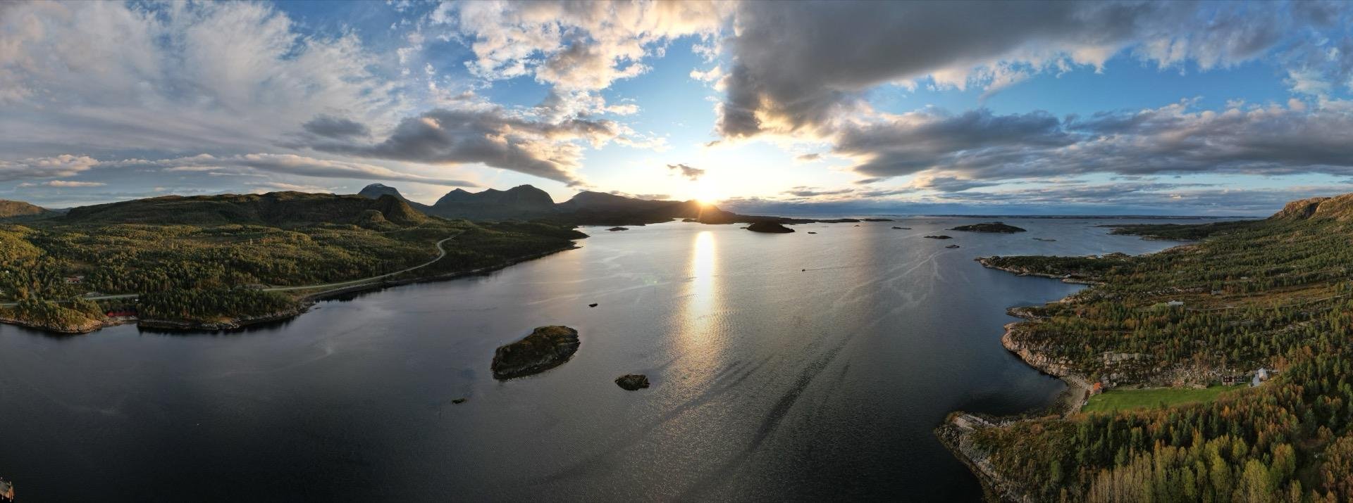 Panoramic view of a serene lake at sunset, flanked by forested hills and scattered islands, under a partly cloudy sky with the sun low on the horizon casting a golden reflection on the water.