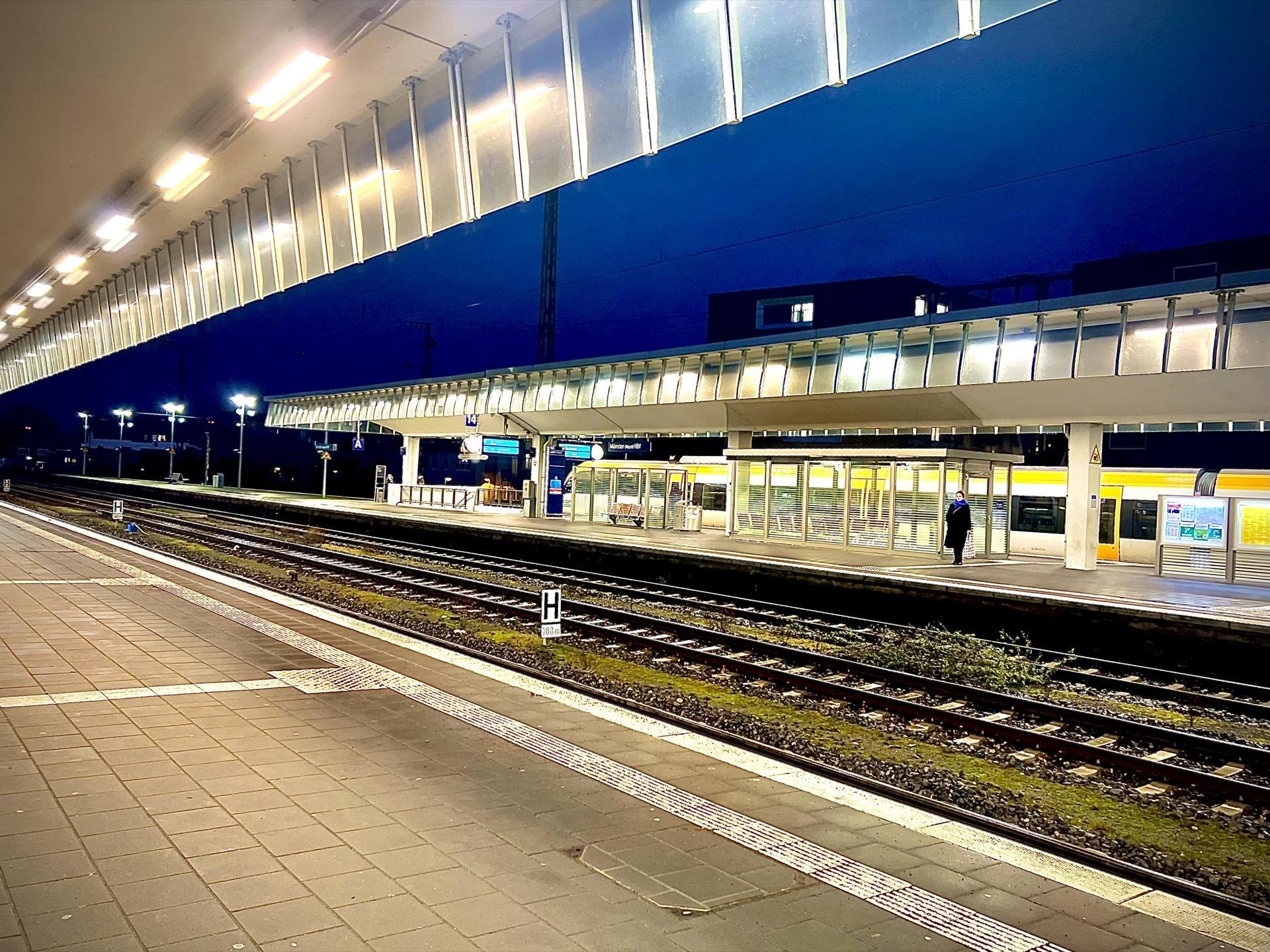 Empty train station platform at night, illuminated by overhead lights, with multiple empty tracks in the foreground and a row of closed waiting areas visible. The station has a modern design with a prominent overhang and minimal pedestrian activity, highlighted by fluorescent lighting along the platform edge.