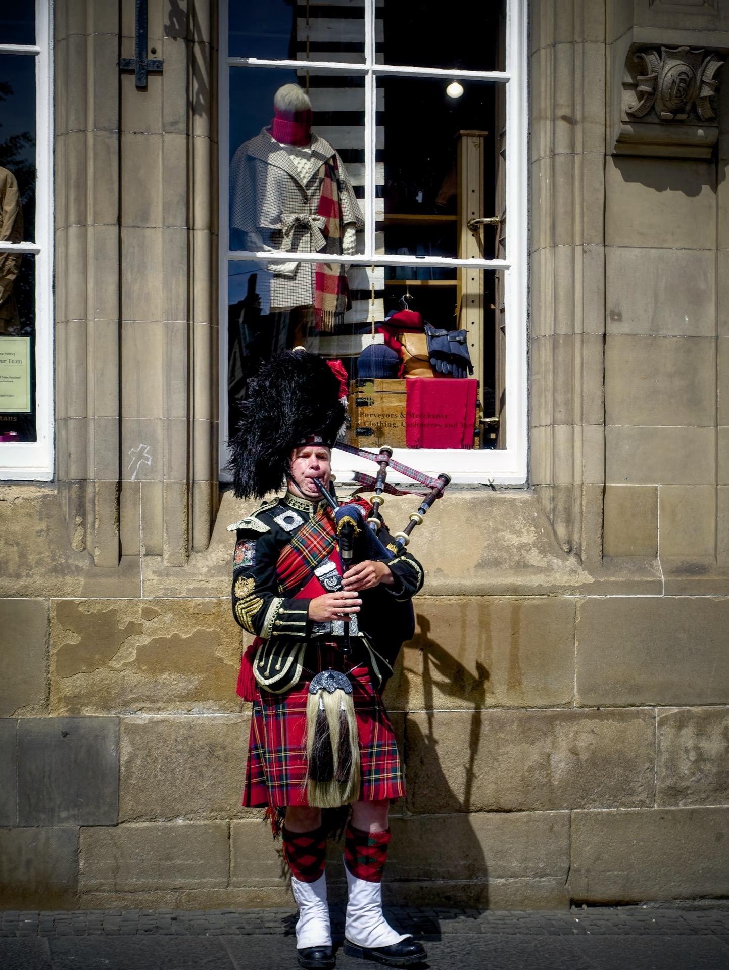 A Scottish bagpiper in traditional dress, including a tartan kilt, sporran, and feathered hat, plays bagpipes on a sunny day beside a building with classical architecture. Behind him, a shop window displays various scarves and draped mannequins. His shadow is visible on the building wall.
