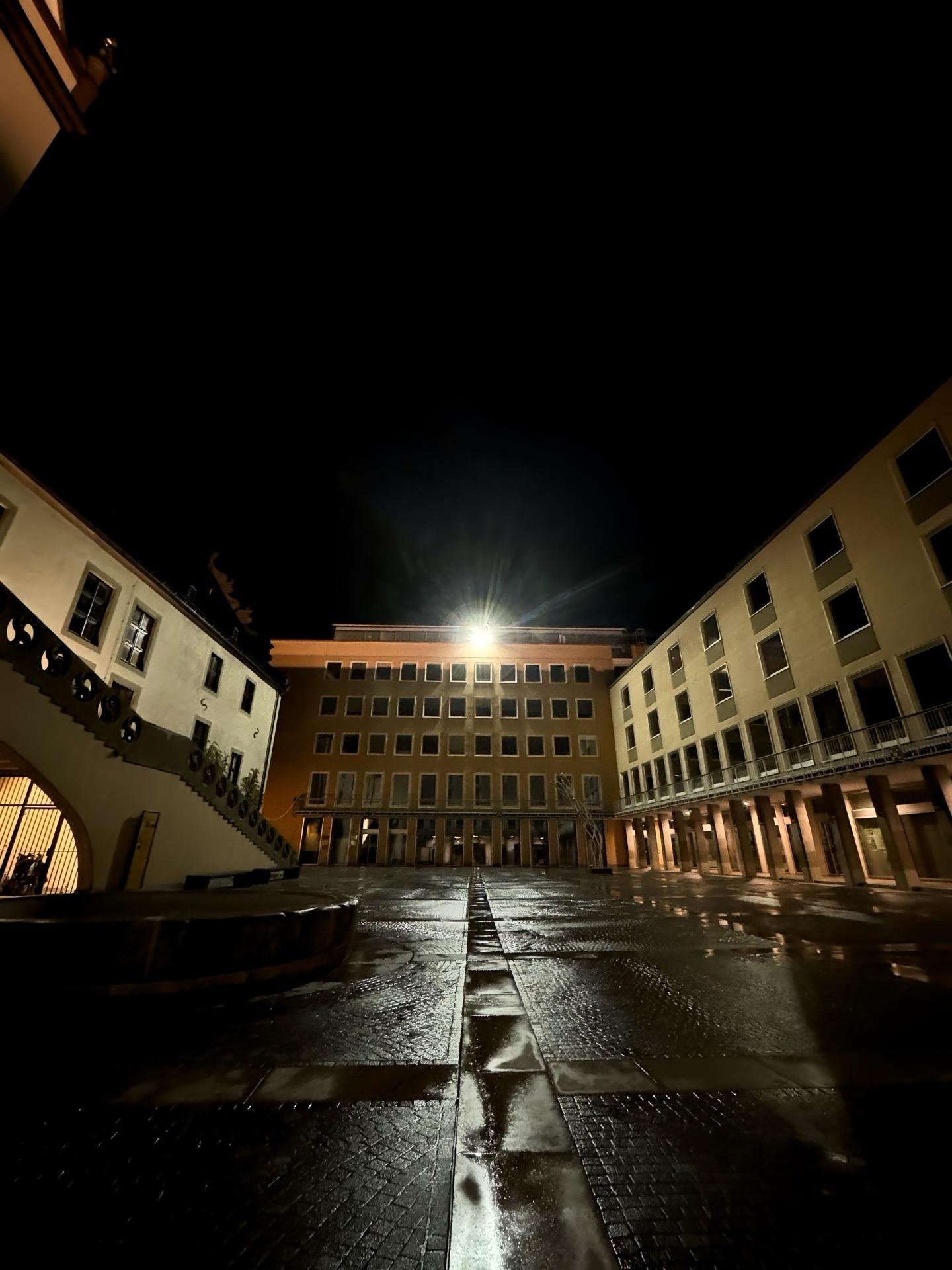 Night view of a wet square flanked by buildings with arches and columns, featuring a bright streetlamp above and a dark sky. A linear pattern on the ground leads directly to the lamp, emphasizing its glow in the scene.