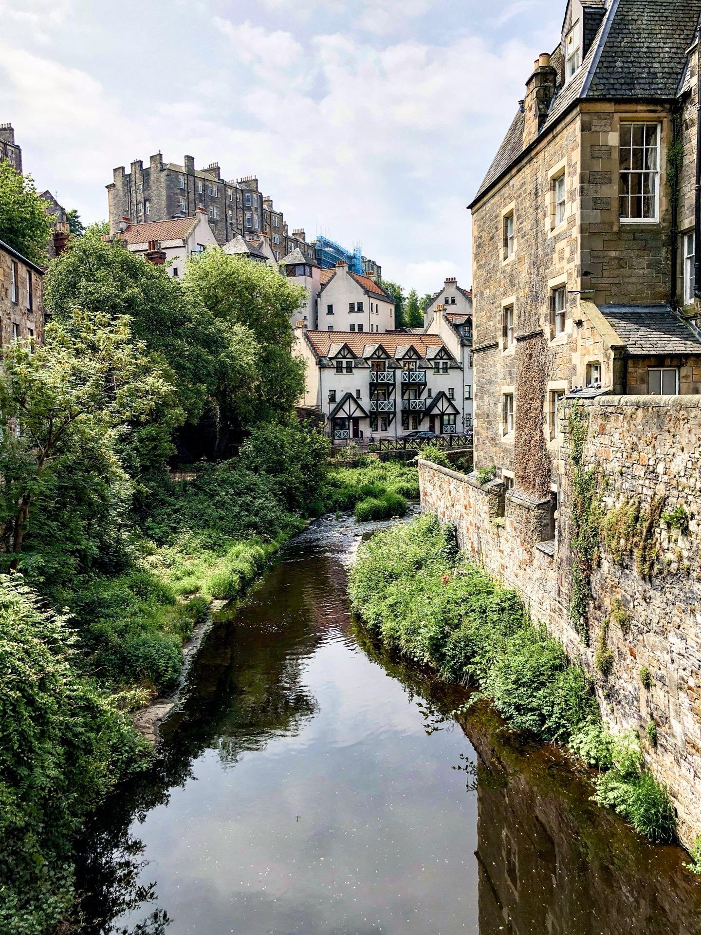 A narrow river runs through a lush, green landscape with an array of historic stone buildings with varying architectural styles on either bank. The foreground shows the river edged by stone walls, beneath which vegetation grows. On the left bank, a Tudor-style building with black and white timber framing rises among other older stone structures. In the background, taller buildings extend up a hill, leading to a visible castle-like structure at the top under a cloudy sky.