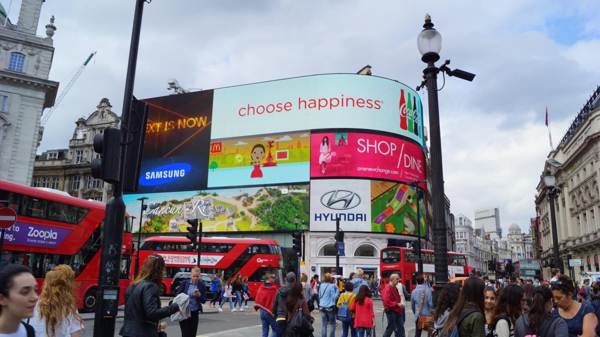 Large, vibrant advertising screens at a bustling urban intersection with pedestrians and double-decker buses, set against a backdrop of historic architecture under a cloudy sky.