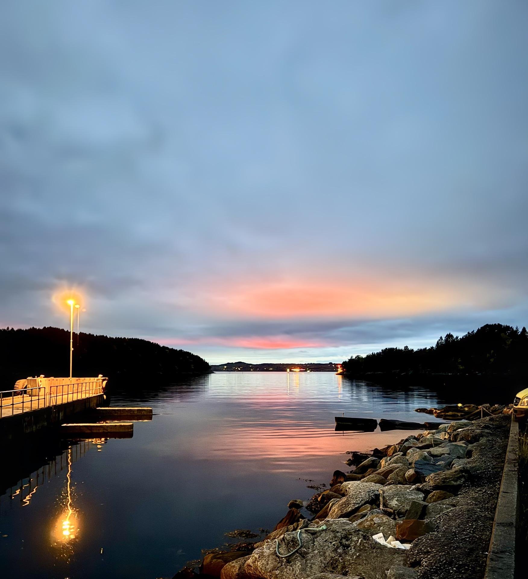 Evening view of a calm water body with a vibrant sunset reflecting off the surface. A lit lamp post stands on a small pier to the left, casting a reflection on the water. Rocky shoreline visible in the foreground with scattered debris and a boat submerged near the edge. The sky gradients from a deep blue to a fiery orange near the horizon.