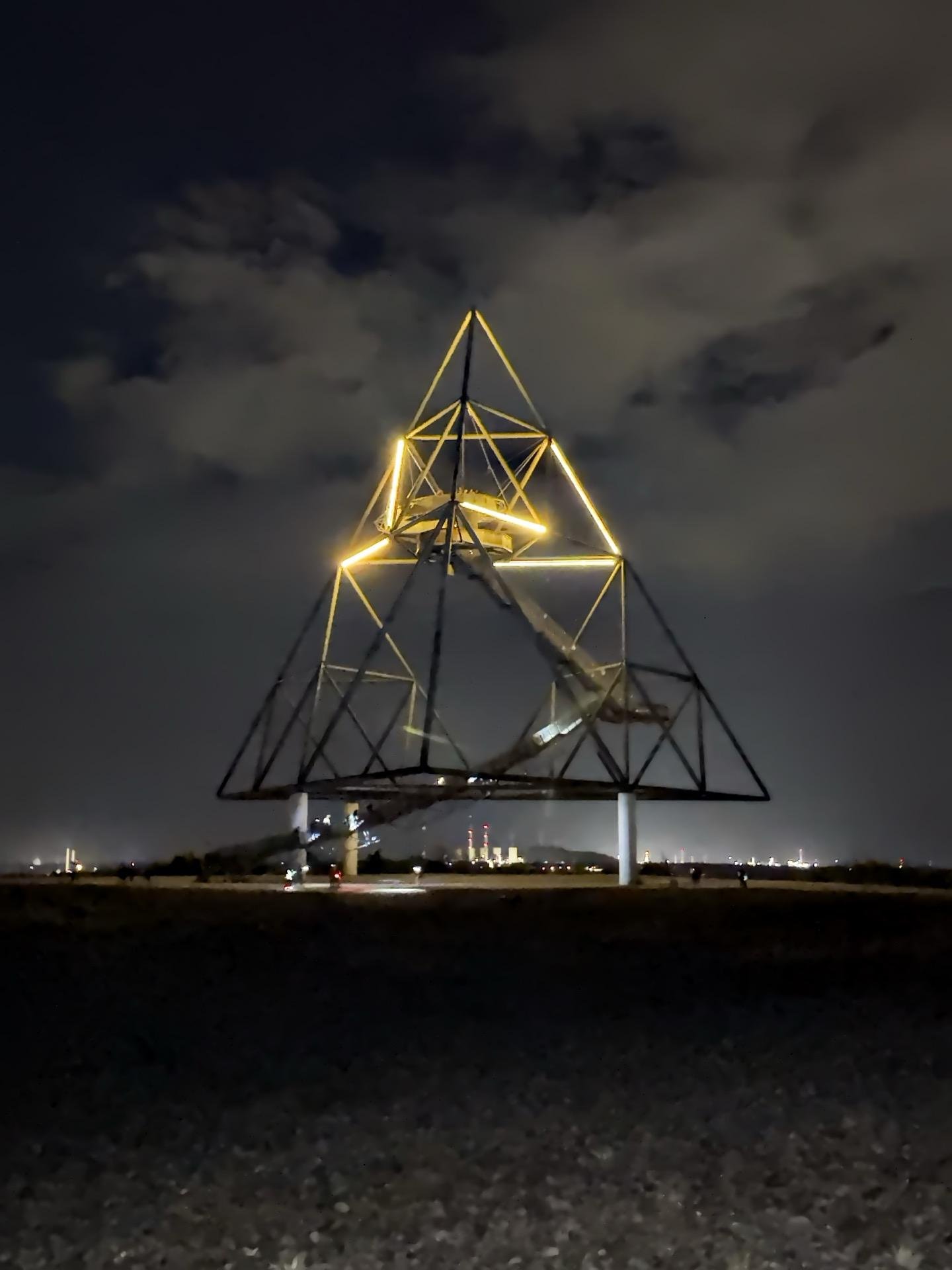 Nighttime photo of a large, illuminated, geometric sculpture made of metal beams, shaped like a pyramid, set against a dark sky with scattered clouds. The landscape shows rough terrain in the foreground and a soft glow of distant lights possibly from a city or industrial area.
