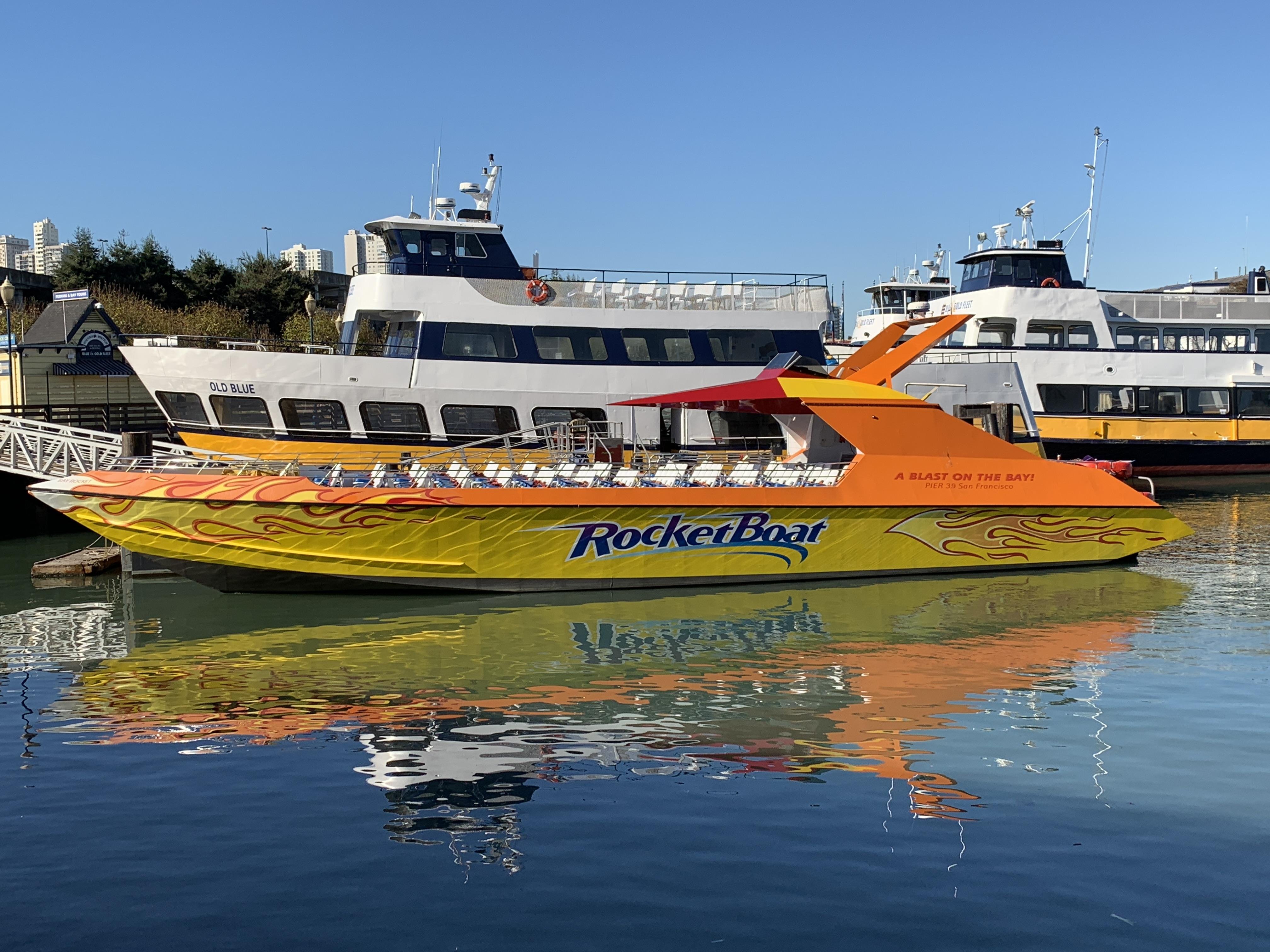 Bright yellow and orange speedboat named "RocketBoat" docked in calm blue water, with vivid reflections visible. The boat features dynamic flame graphics and is moored next to a grey and blue ferry named "Otis Blue." Clear sky and distant city skyline in the background.