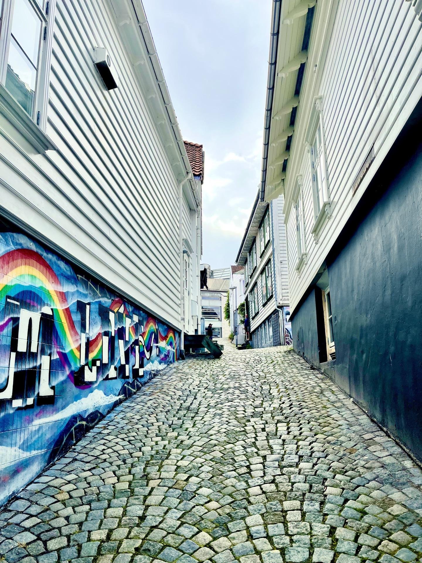 Narrow cobblestone street flanked by traditional white wooden houses with rainbows painted on the left wall. One side of the street has houses with exposed vertical siding while the other features houses with shutters and raised foundations. Overcast sky visible above the enclosed, quaint urban path.